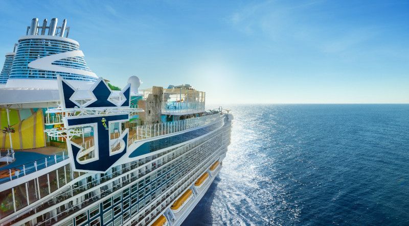 Large cruise ship sailing on a sunny, blue ocean. Royal Caribbean logo visible.