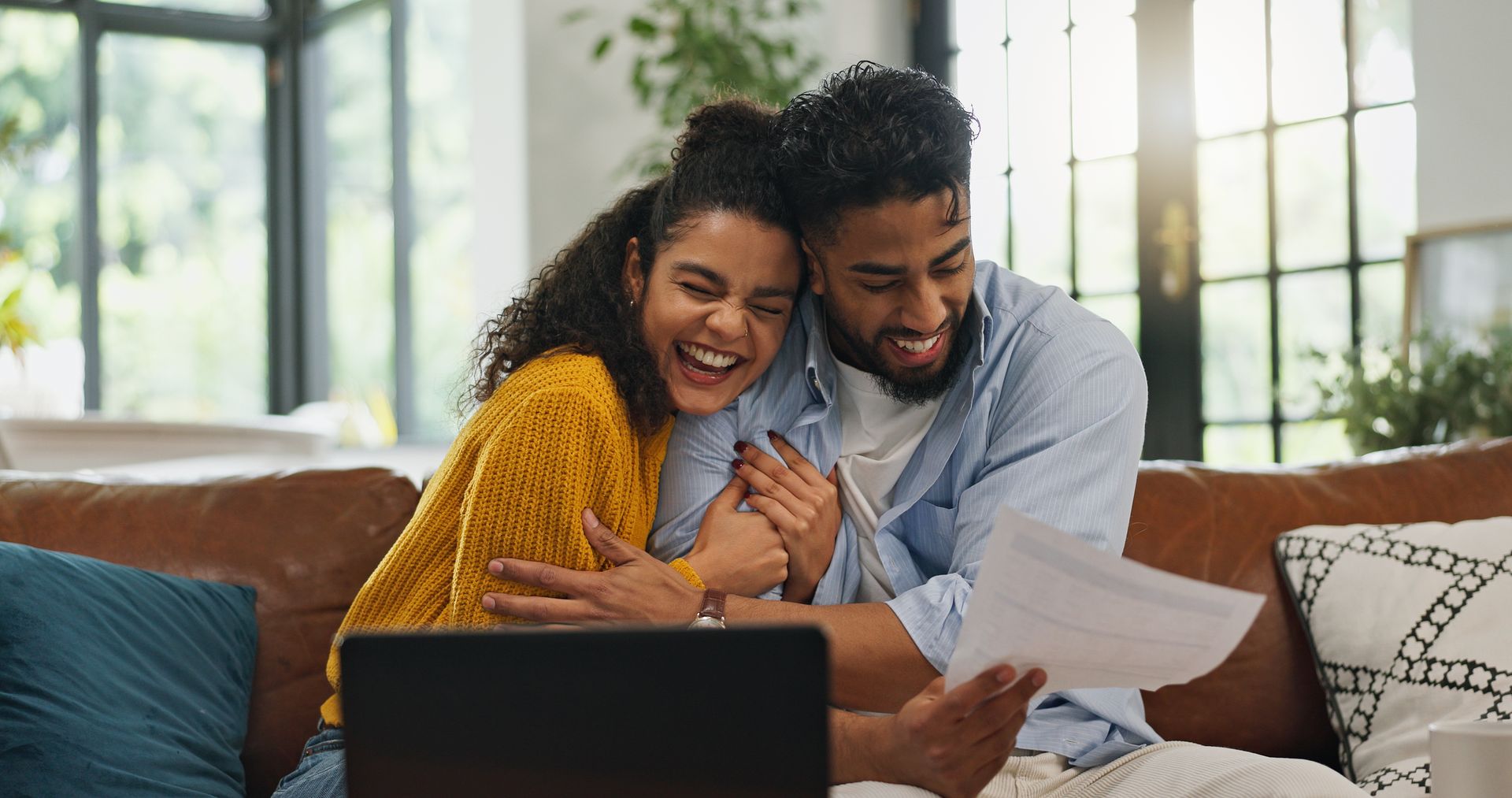 Couple on a couch, laughing and looking at papers, laptop in front of them in a bright living room.