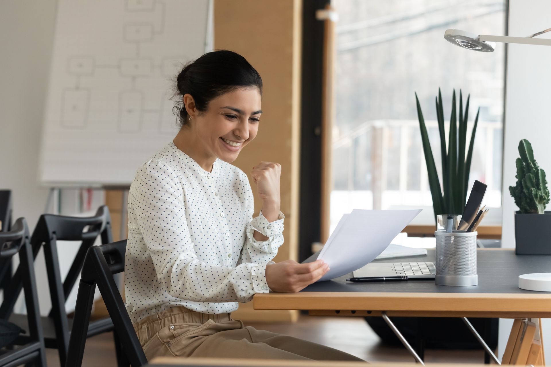 Woman at desk smiles, raising fist, holding document, appearing happy. Office setting with plant. Woman at desk smiles, raising fist, holding document, appearing happy. Office setting with plant.