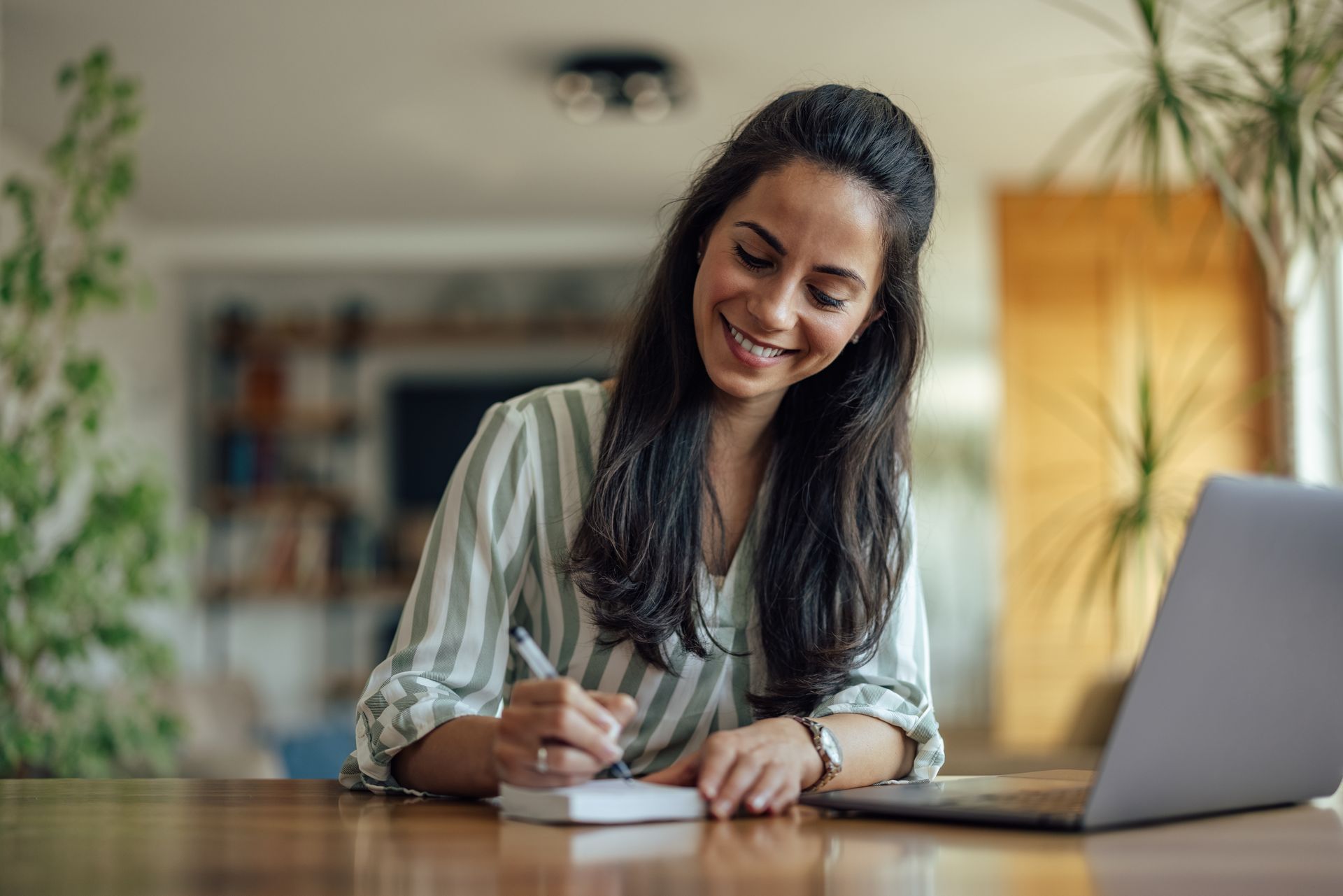 Woman writing in notebook, smiling, with laptop on table. Interior with plants and natural light. Woman writing in notebook, smiling, with laptop on table. Interior with plants and natural light.