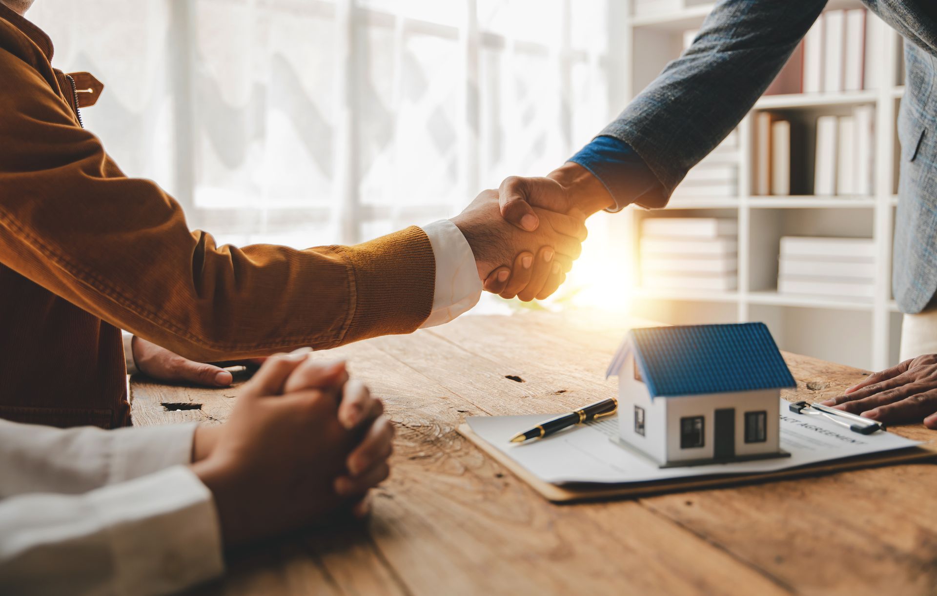 People shaking hands over a miniature house, contract, and pen on a wooden table. Sunlight streams in. People shaking hands over a miniature house, contract, and pen on a wooden table. Sunlight streams in.