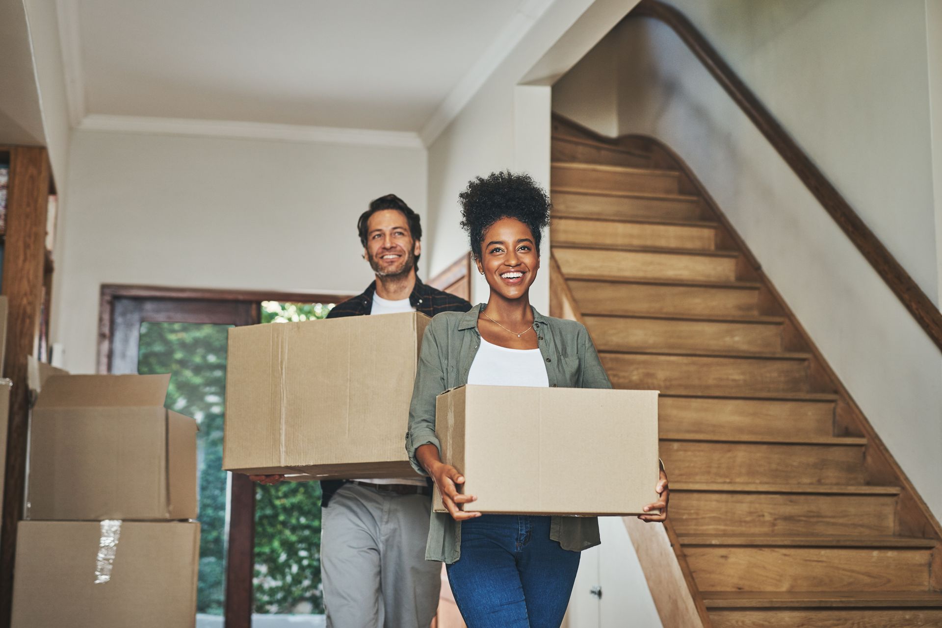 Couple carrying moving boxes, smiling, entering a house with stairs.