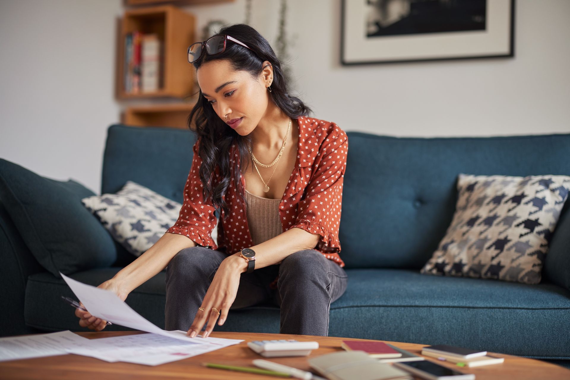 Woman on a blue sofa reviews paperwork, with glasses perched on her head, in a living room.