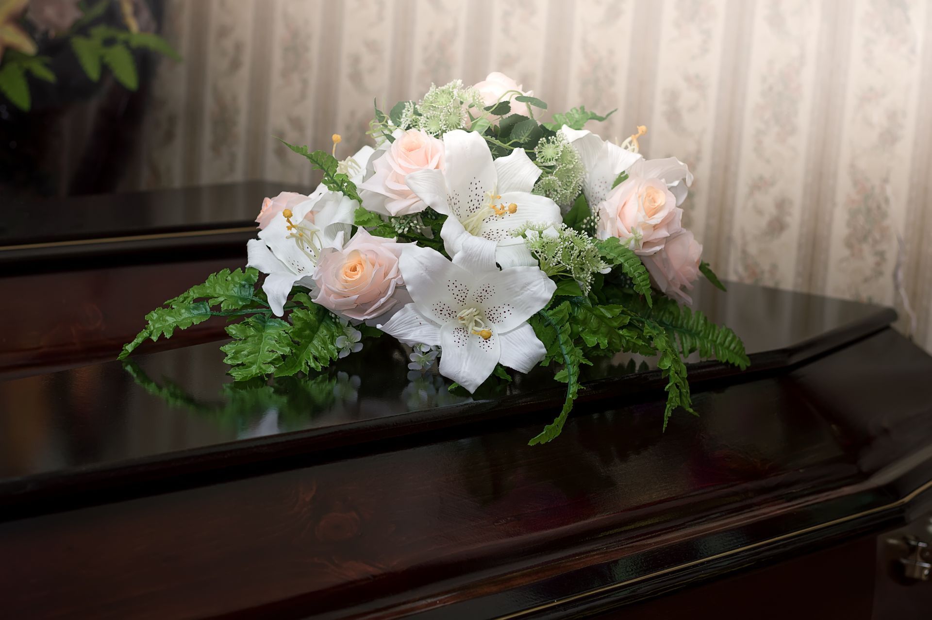 A floral arrangement of white lilies and pale pink roses sits atop a polished dark wood casket.