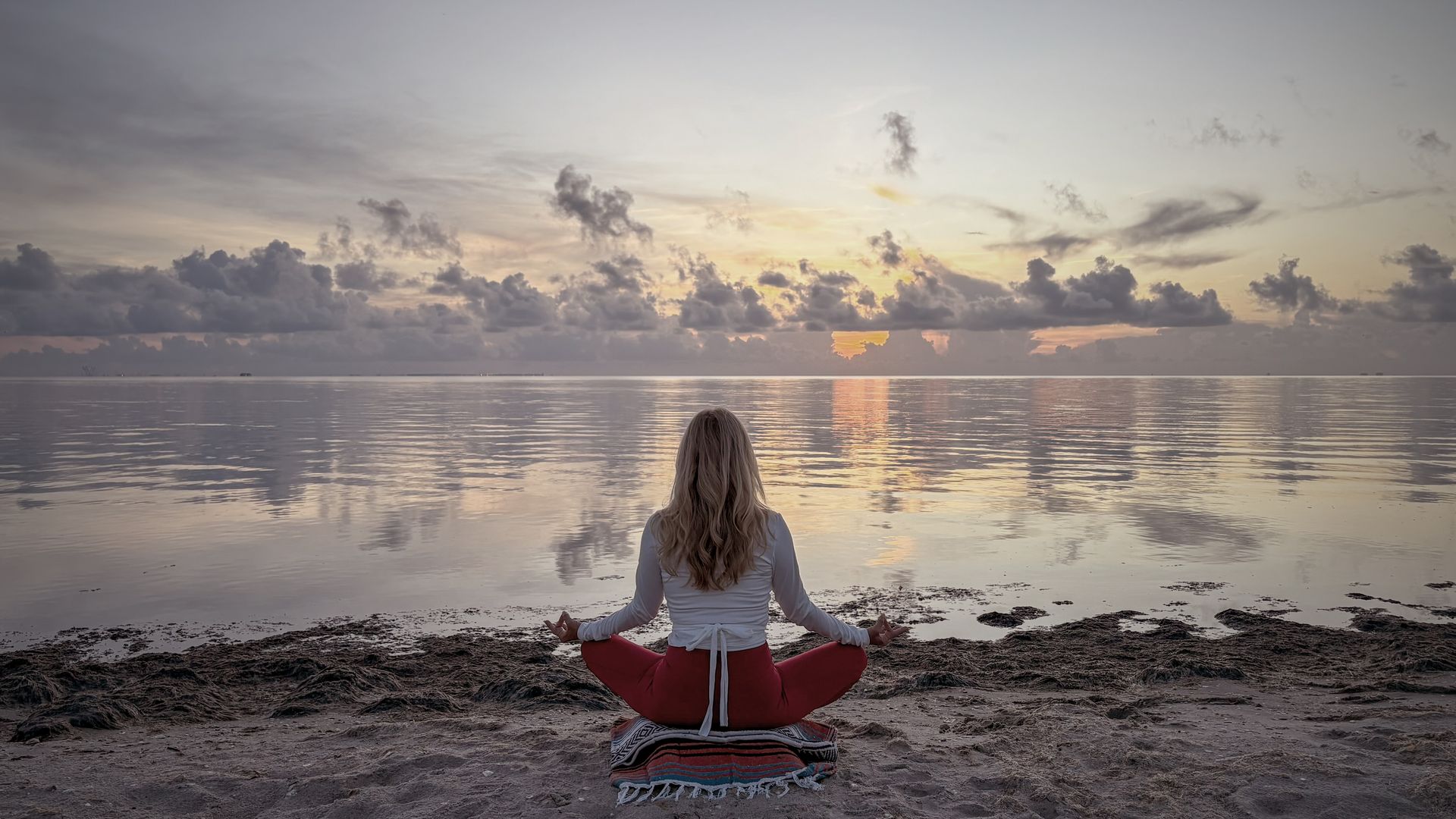 Woman meditating by the water at sunset, with red pants and white shirt.