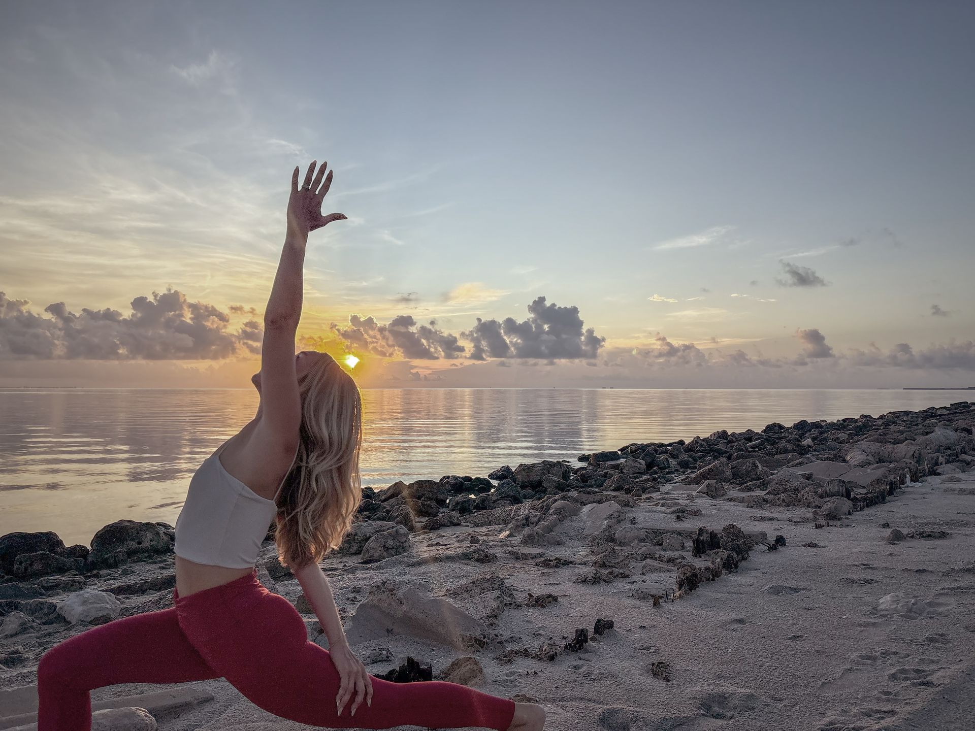 Woman in yoga pose on beach at sunrise, arm raised, red leggings, white top, ocean backdrop.