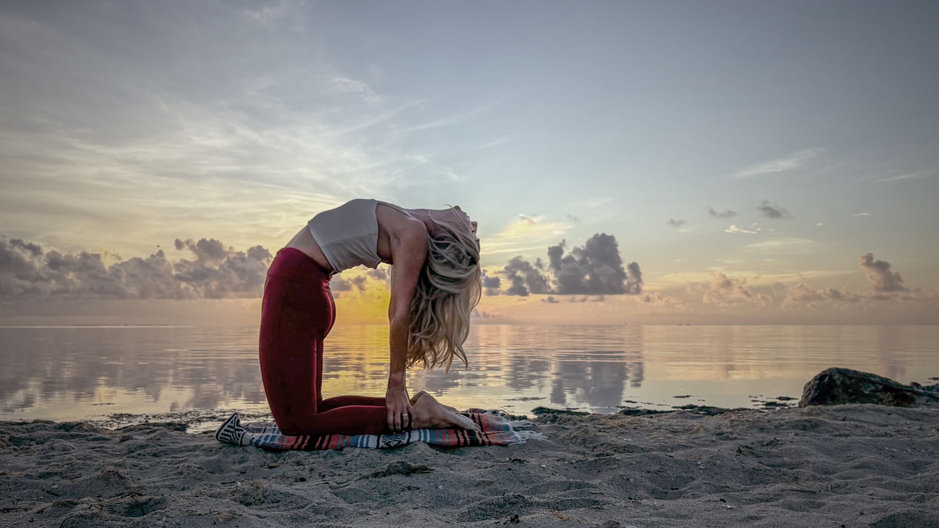 Woman in yoga pose, arched back, on beach at sunset, wearing red leggings, white top.