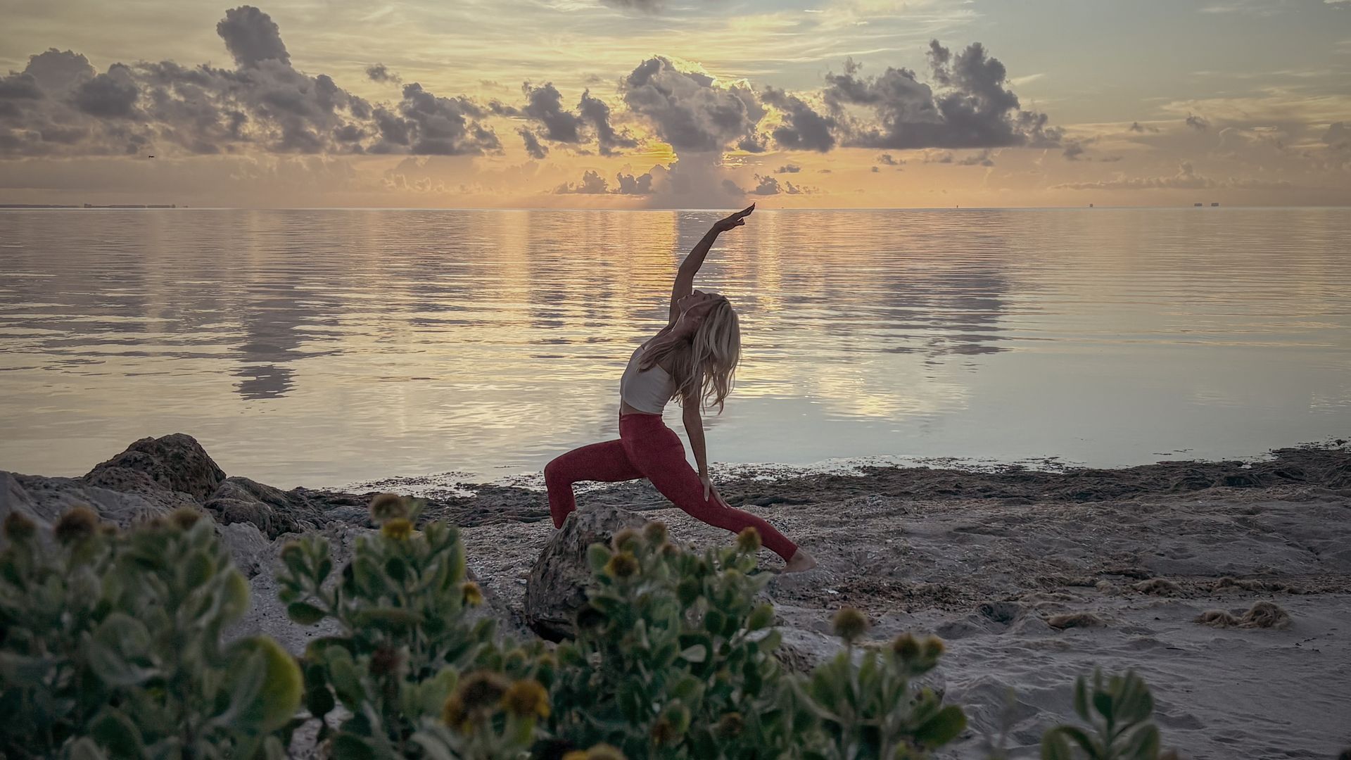 Woman doing yoga on a beach at sunset, wearing red leggings and white top, arm raised.