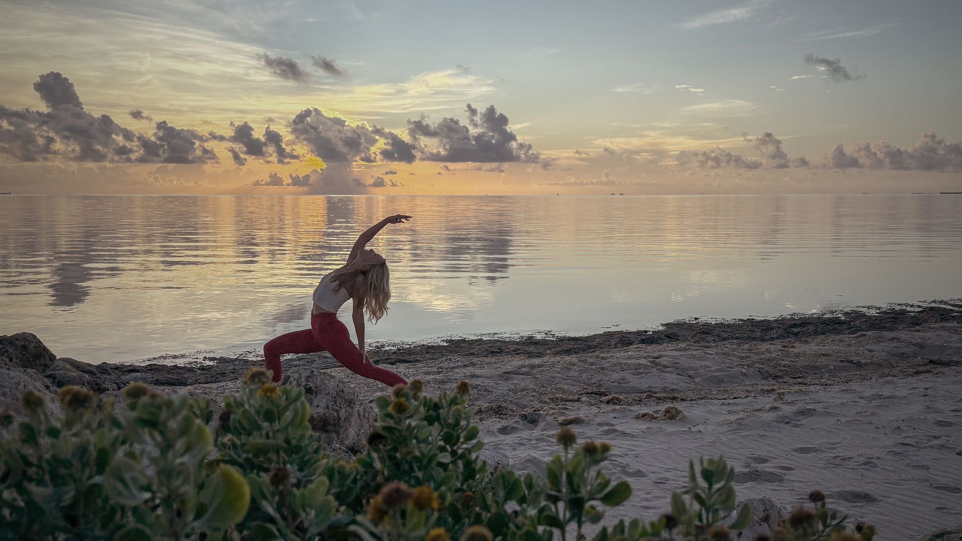 Woman in yoga pose on beach at sunset, wearing red pants, ocean in background.