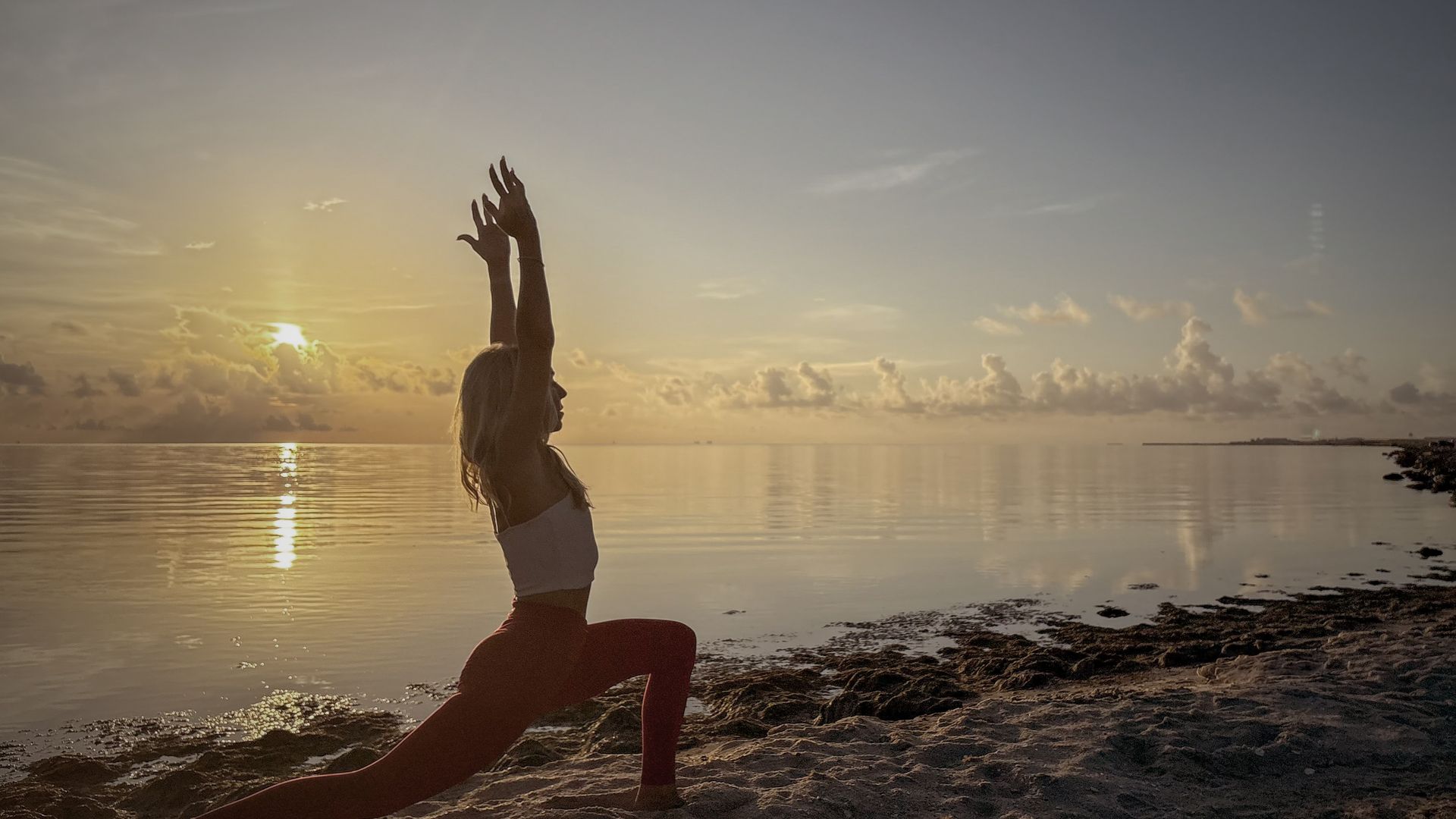 Woman in yoga pose on a beach at sunrise, arms raised, orange leggings, white top, water in background.