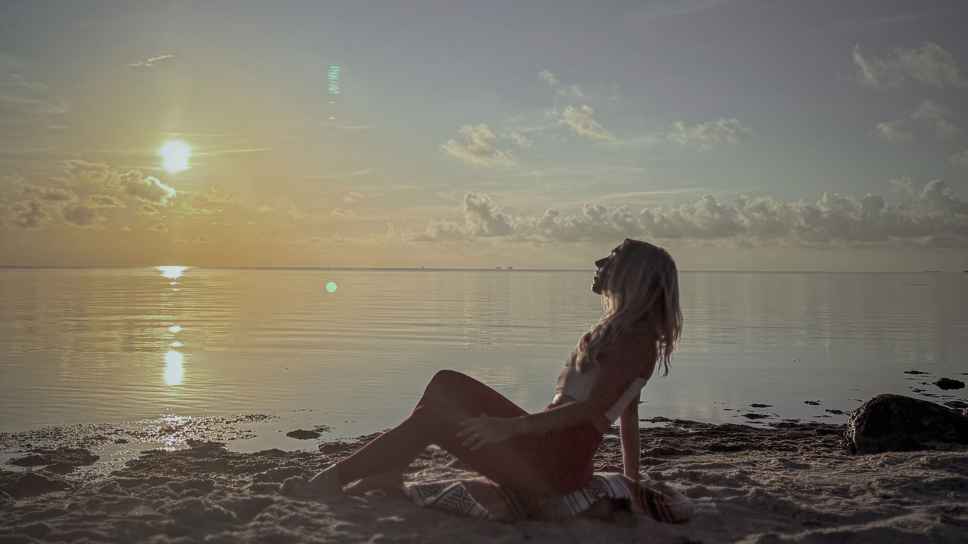 Woman sitting on beach, looking up at sunset over ocean.
