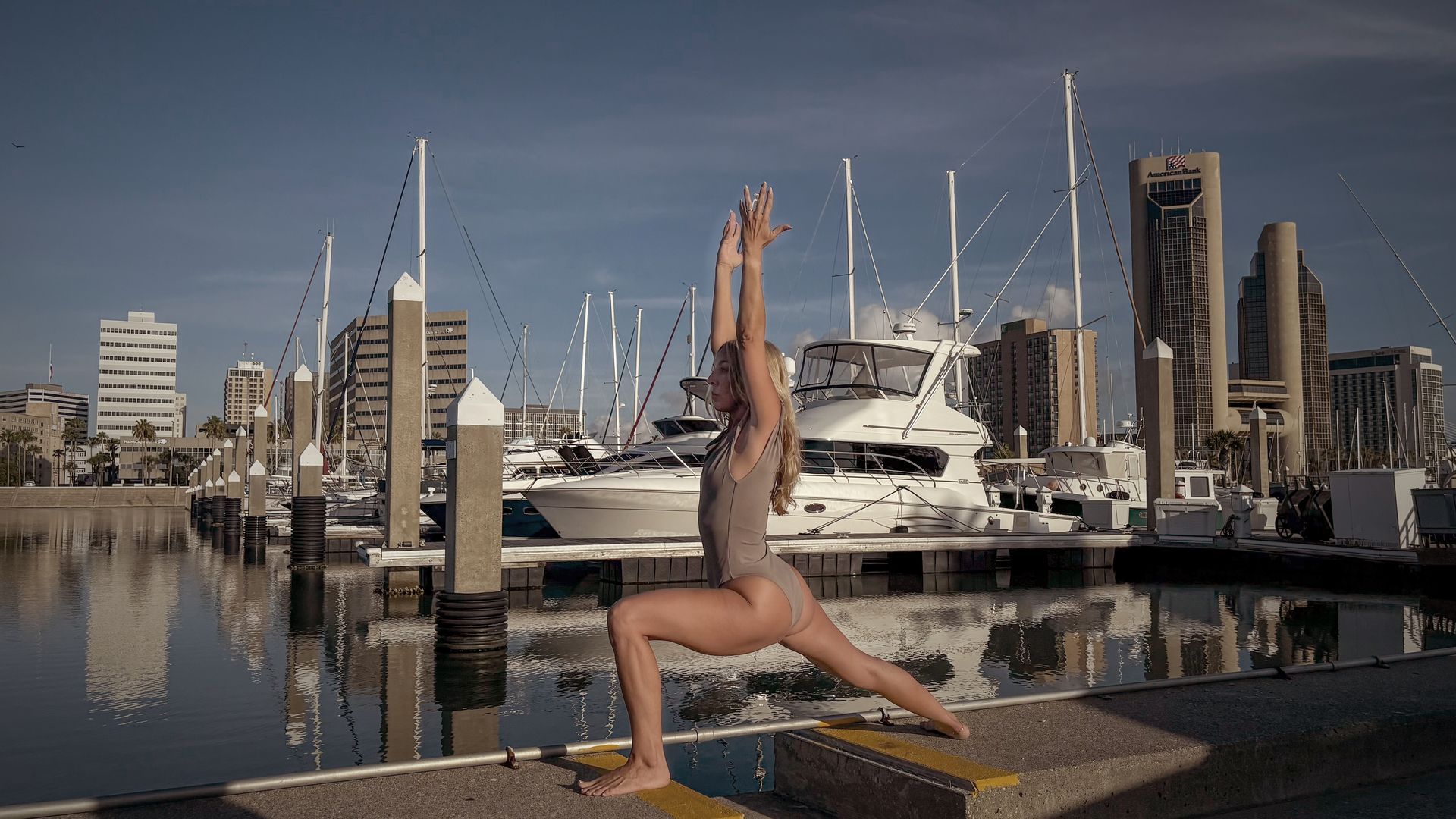 Woman in a yoga pose outdoors by a harbor with boats and buildings.