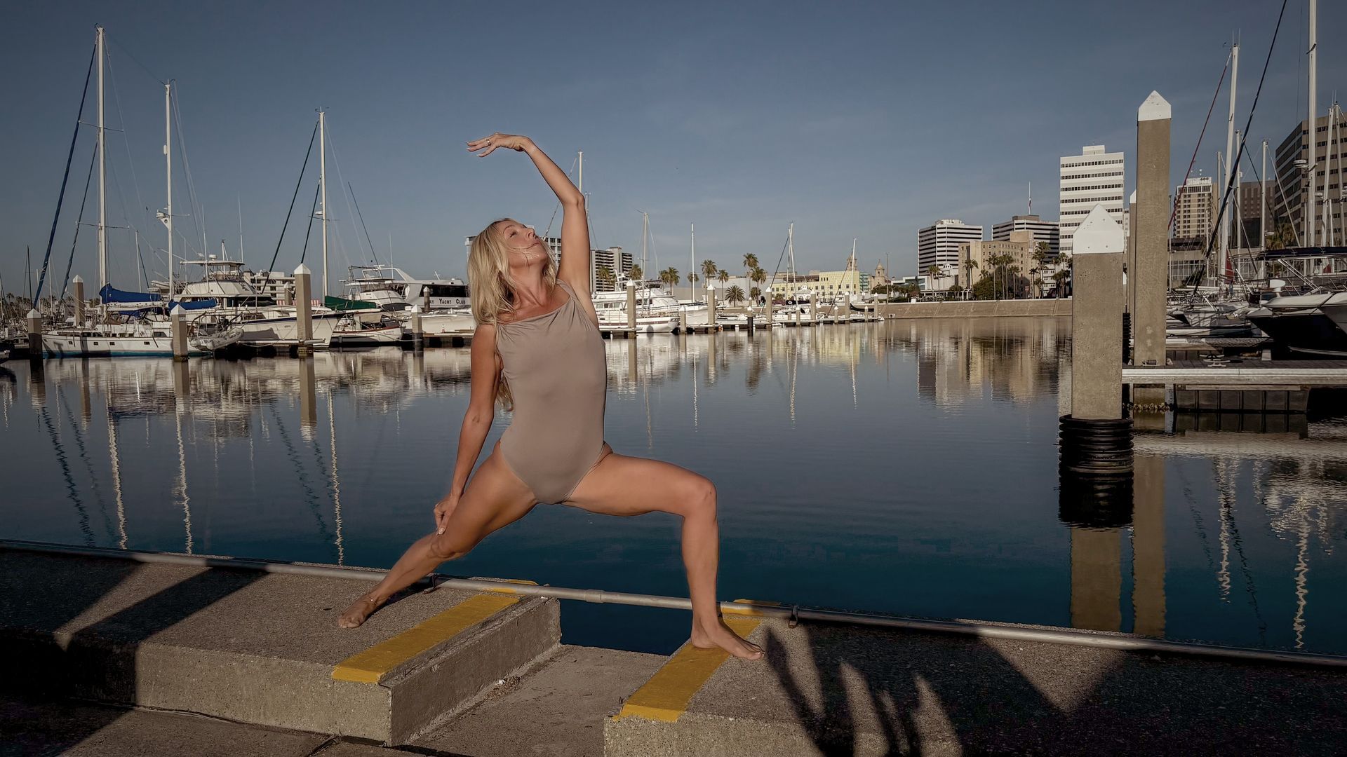 Woman in a yoga pose on a dock, arm raised, harbor and boats in background.