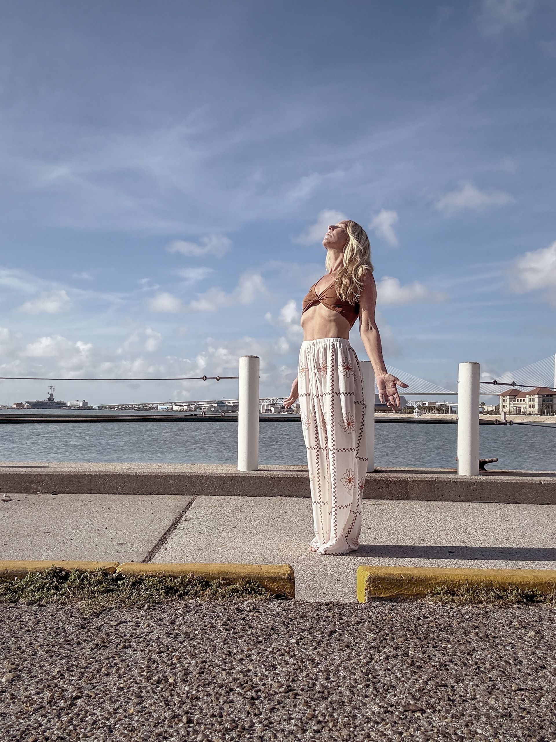 Blonde woman in a brown bikini top and white pants, looking up at the sky near a body of water.