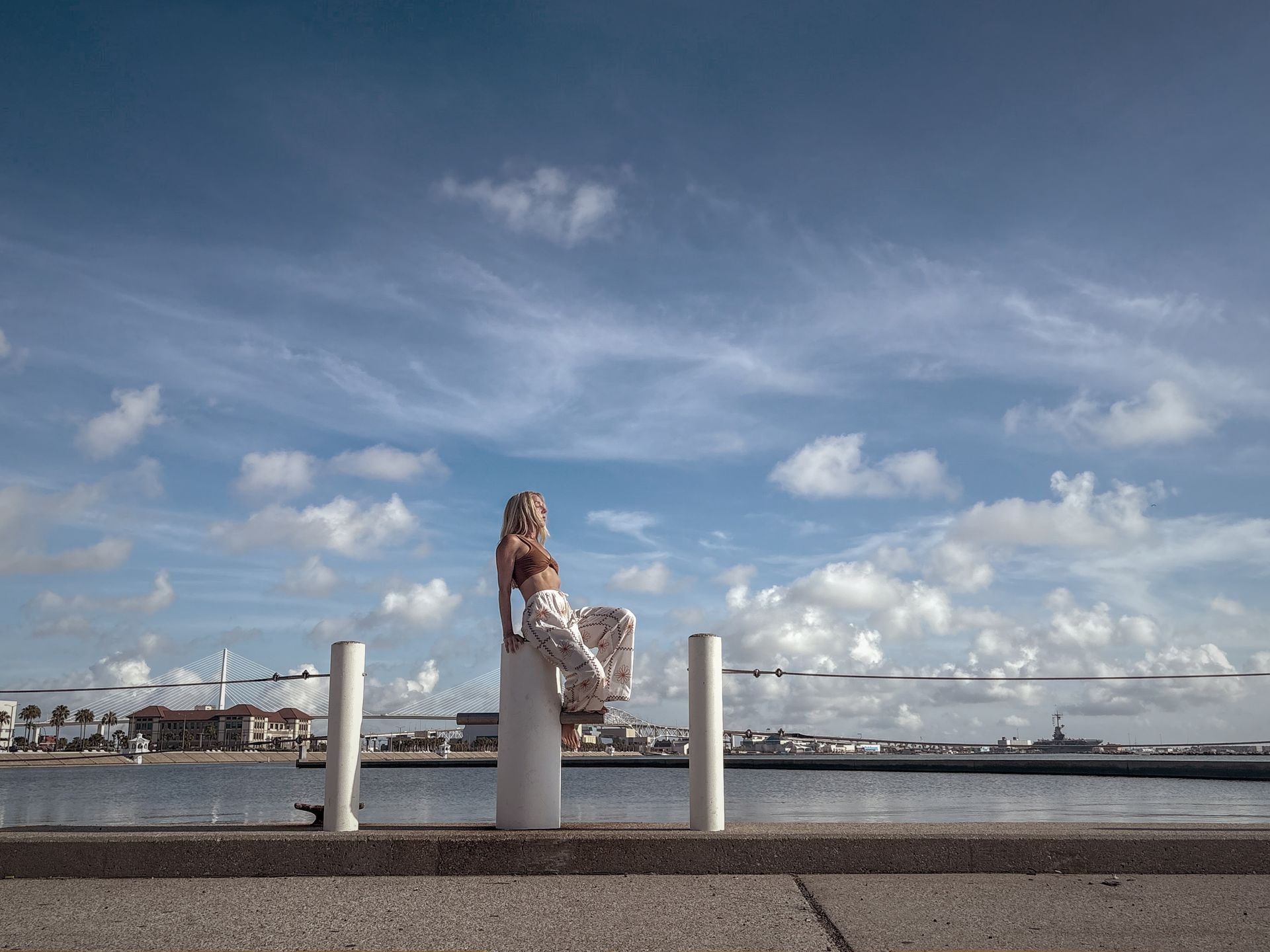 Statue on a white pillar by the water under a blue sky with clouds; buildings in the background.
