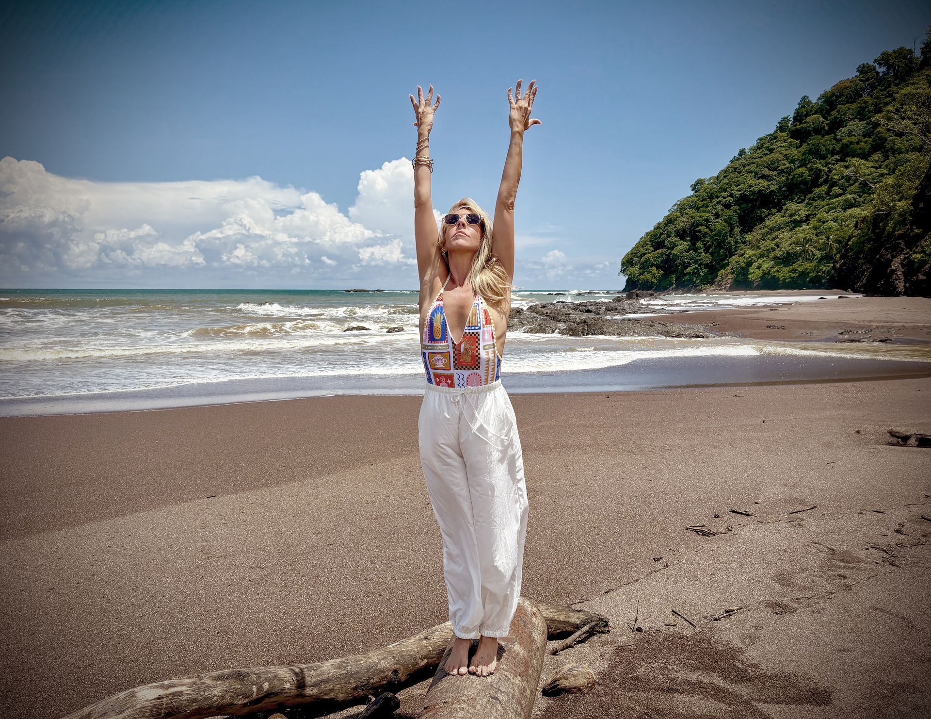 Woman on beach with arms raised, sunny day, ocean and foliage in background.