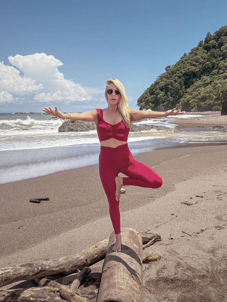 A woman is standing in a yoga pose on a log on the beach.