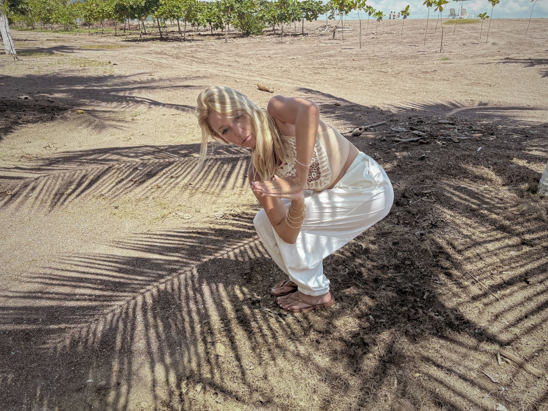 Blonde woman in white pants squats outdoors under palm tree shadows.