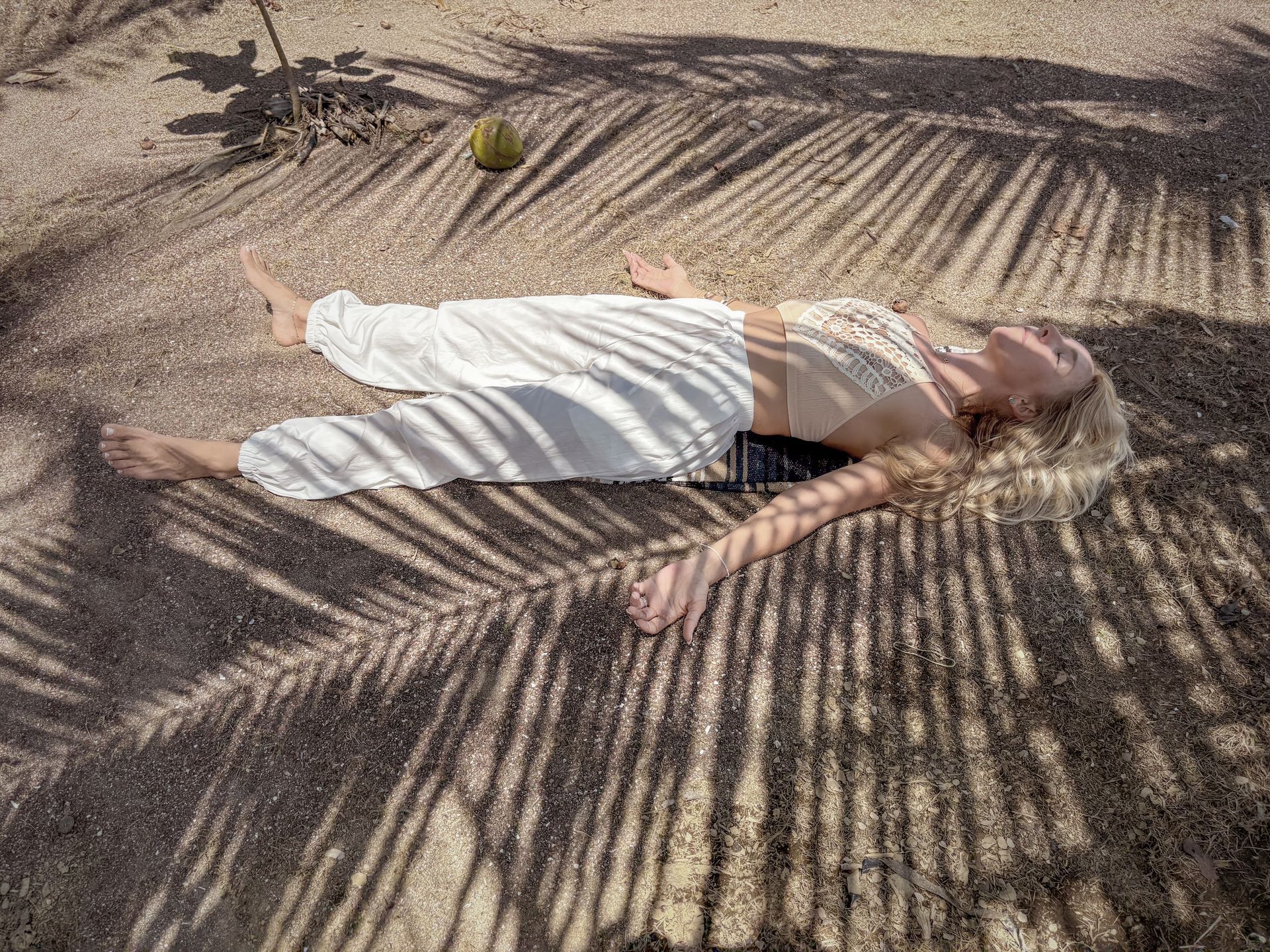 Woman lying on sand under palm tree shadows, wearing white.