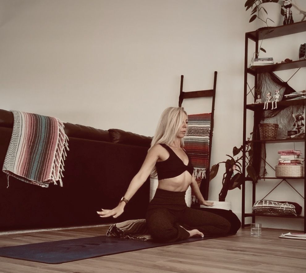 Woman in yoga pose on mat, arms outstretched. Interior setting with bookshelf, couch, and wooden floor.