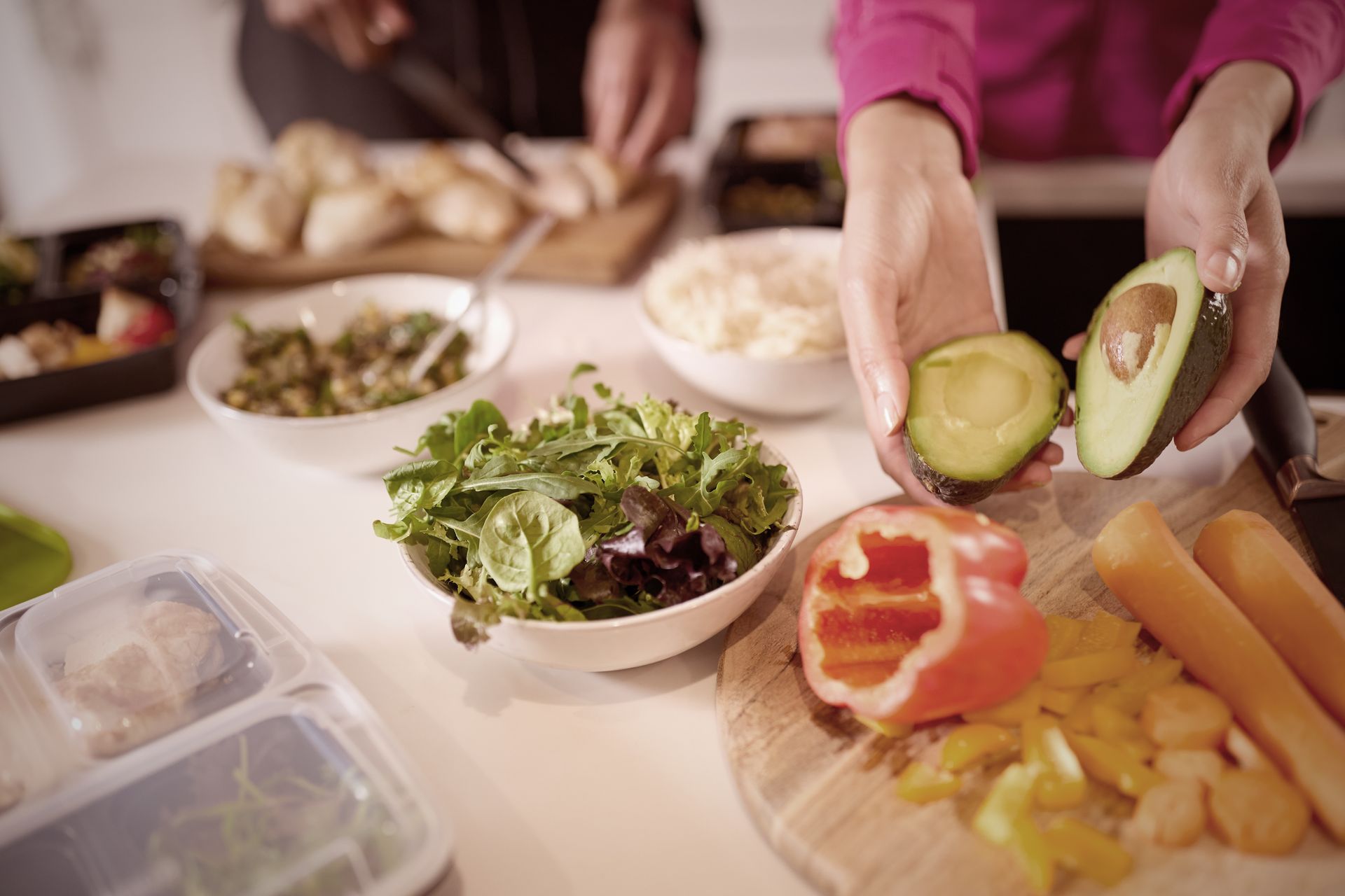 Hands holding avocado halves, surrounded by salad, cut vegetables, and prepared food on a white countertop.
