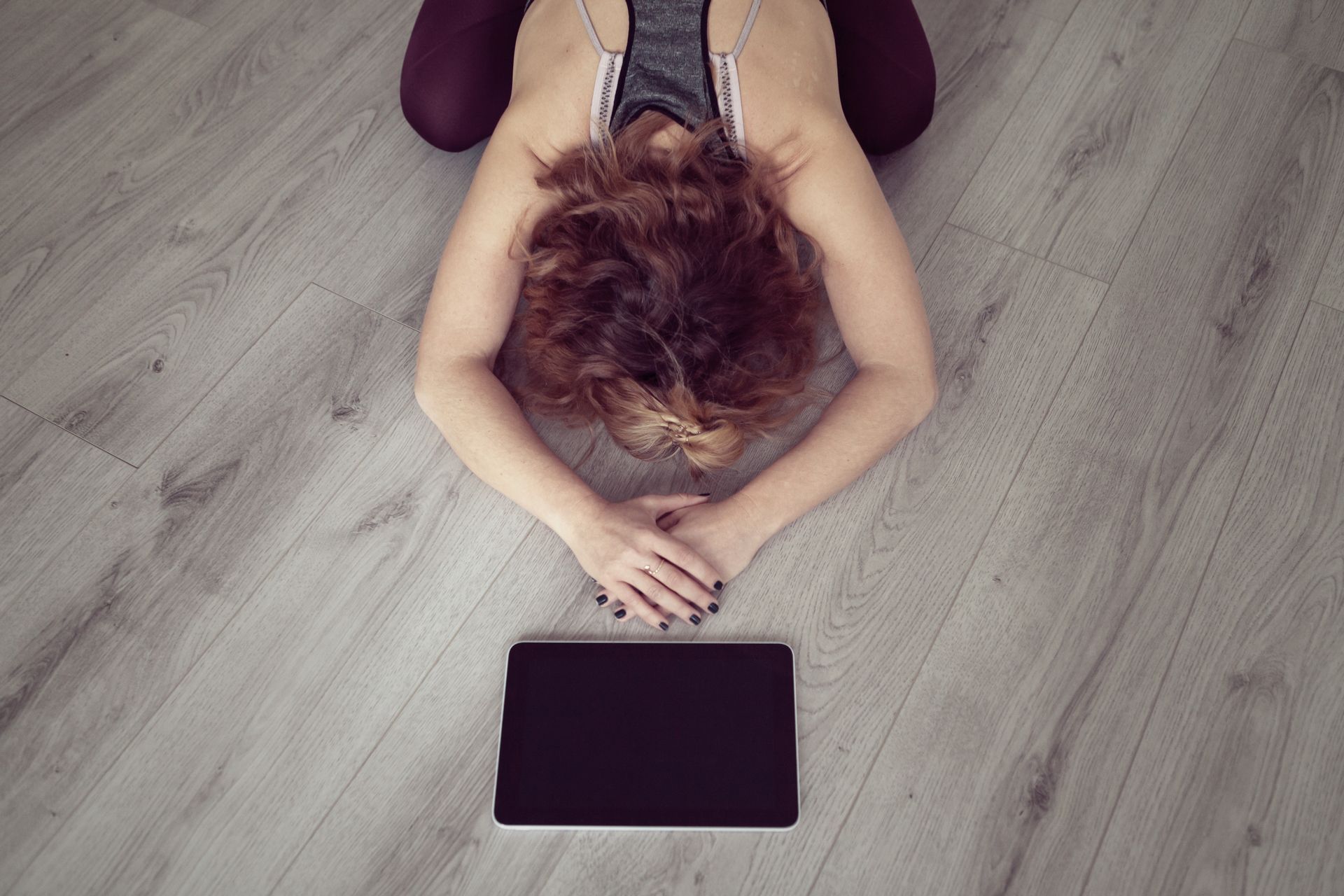 Woman in yoga pose, head down, hands near a tablet on a wooden floor.