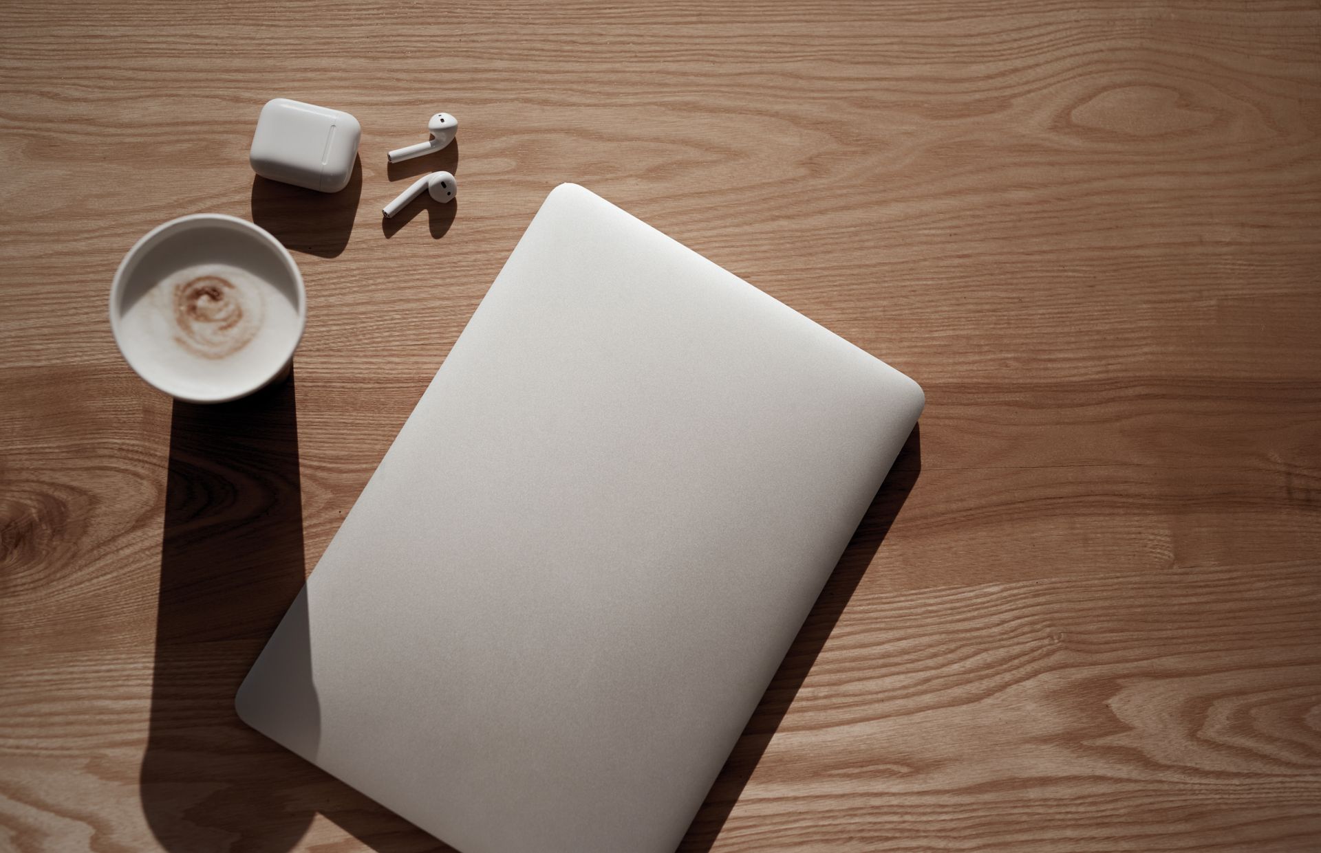 Closed laptop, coffee, earbuds, and charging case on a wooden surface.