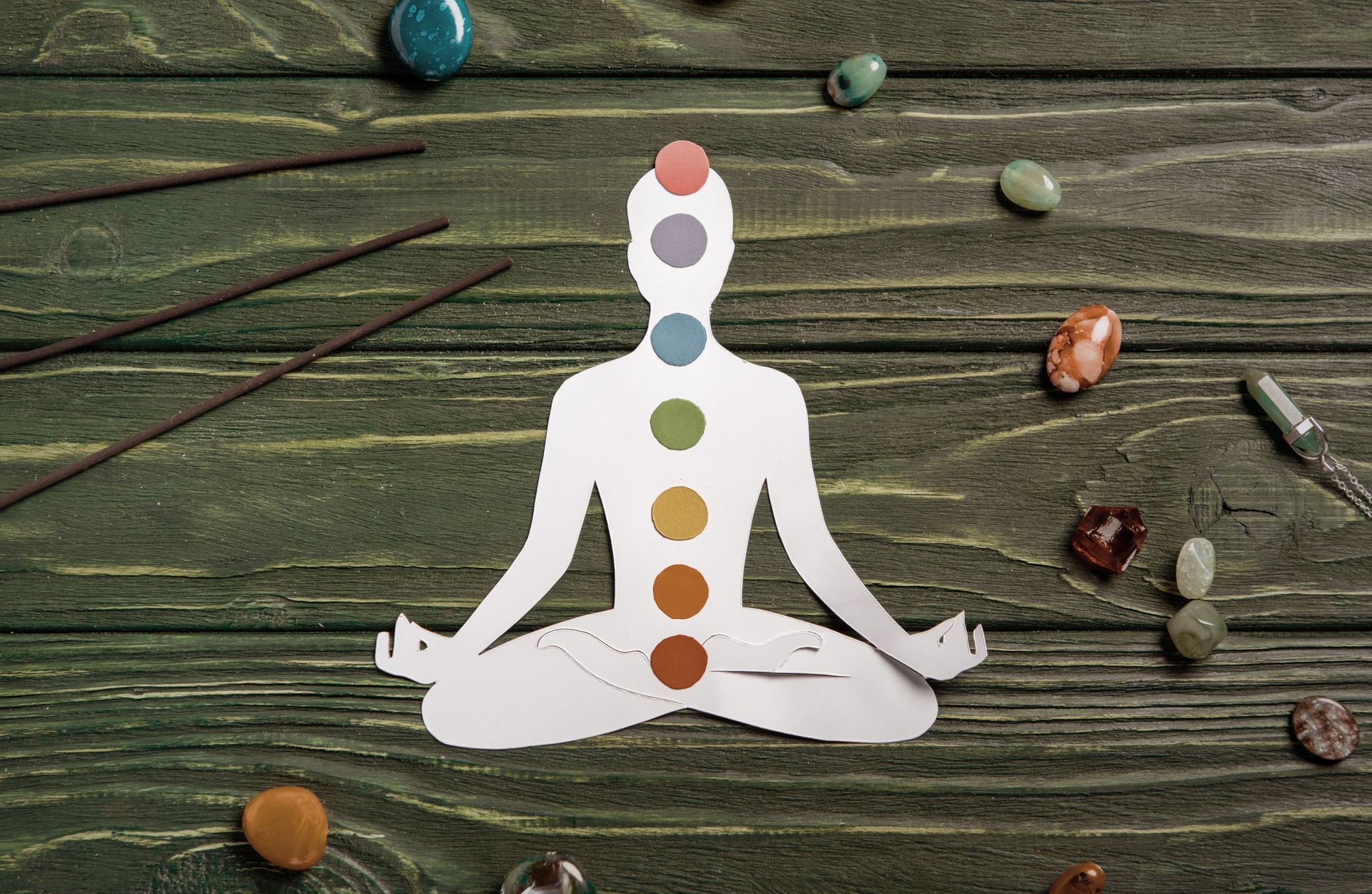 Paper cutout of a person in the lotus position with chakras highlighted, surrounded by crystals and incense on a wooden surface.