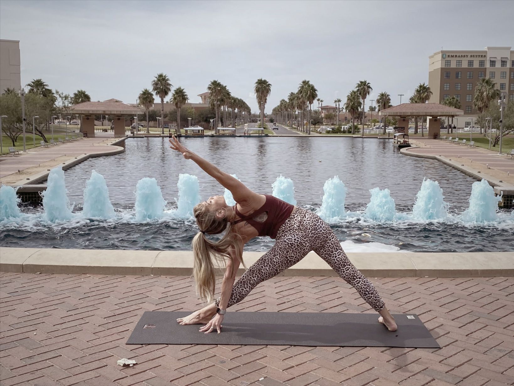 Woman in yoga pose outdoors by a fountain, reaching up with arm.