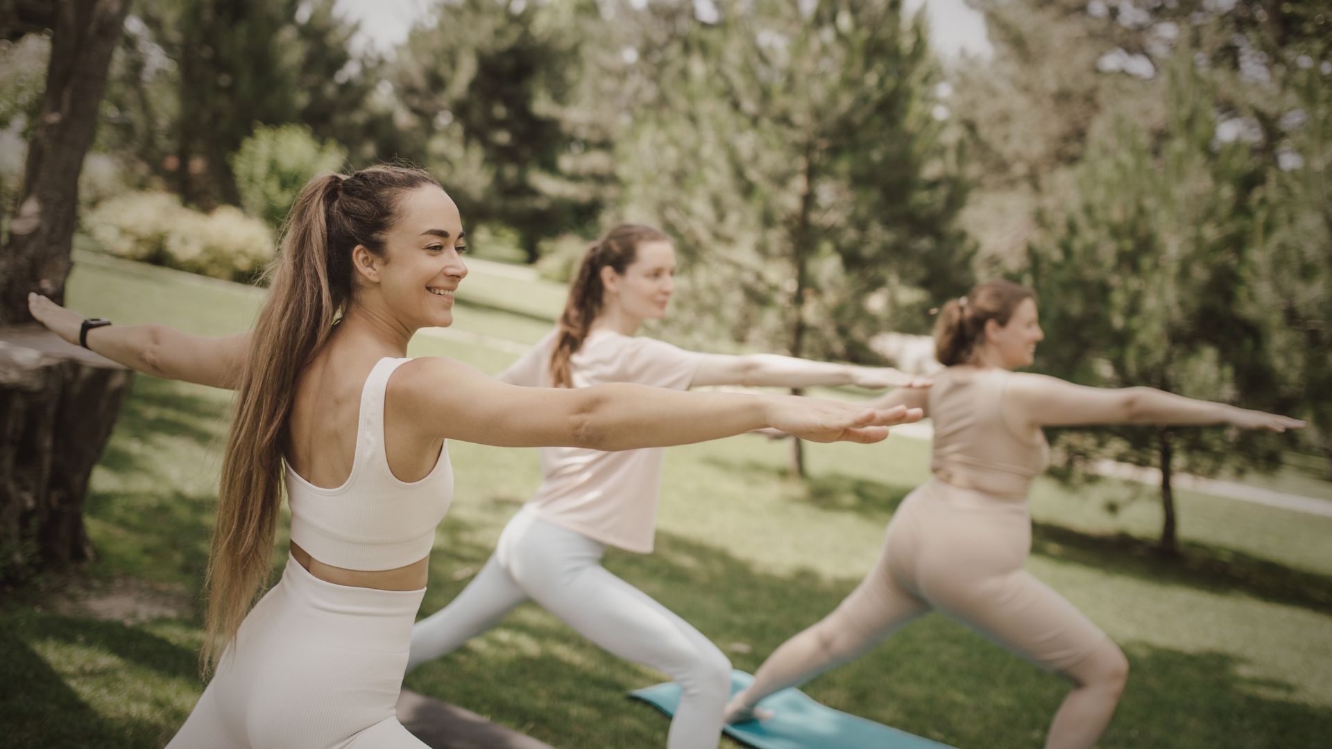 Three women in athletic wear practicing yoga outdoors. They are extending their arms in a pose.