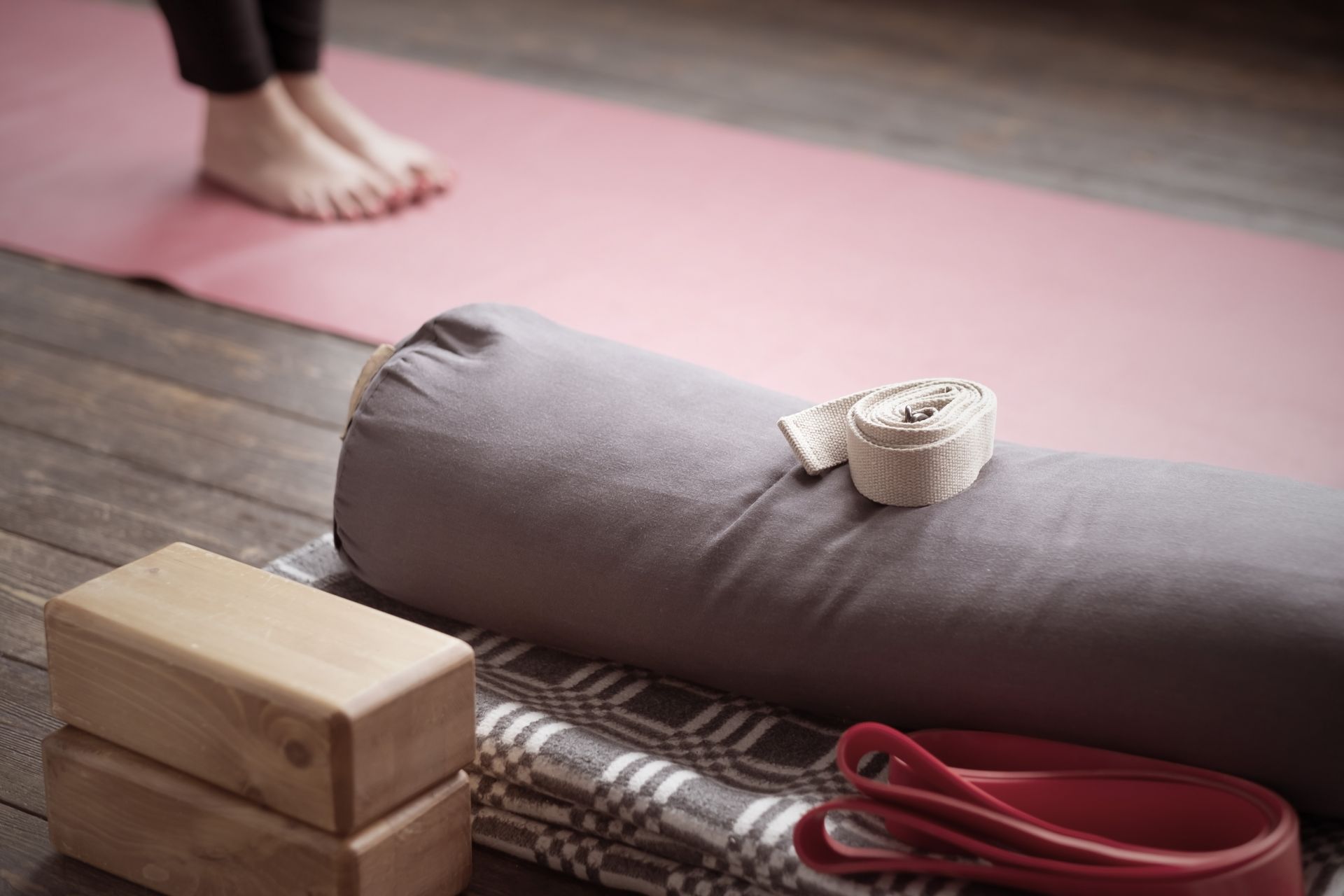 Yoga props: blocks, bolster, strap, and mat on a wooden floor. Bare feet in background.