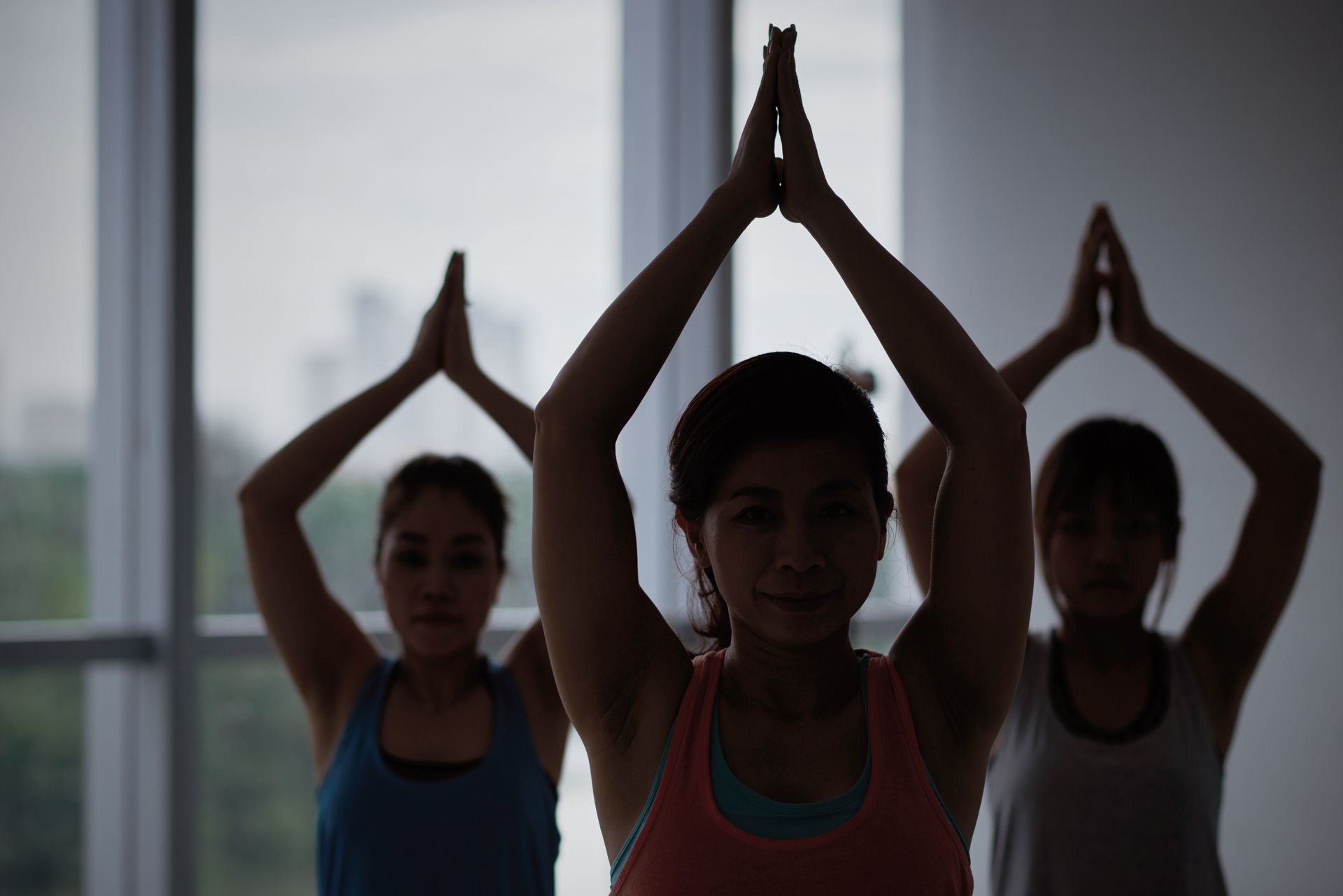 Three women doing yoga, arms overhead in prayer pose, by a window.