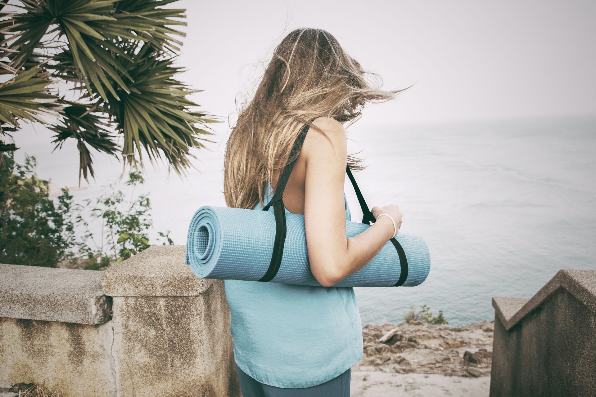 Woman with long hair carries a rolled blue yoga mat over her shoulder near the ocean.