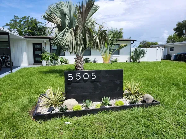 Black house number sign in a landscaped yard; 5505 on black rectangle.