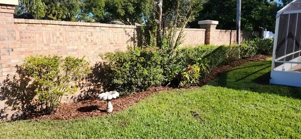 Green lawn bordered by brick wall, shrubbery, and mulch, with bird bath and screen enclosure.