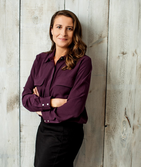 A woman in a purple shirt and black skirt is standing in front of a wooden wall.