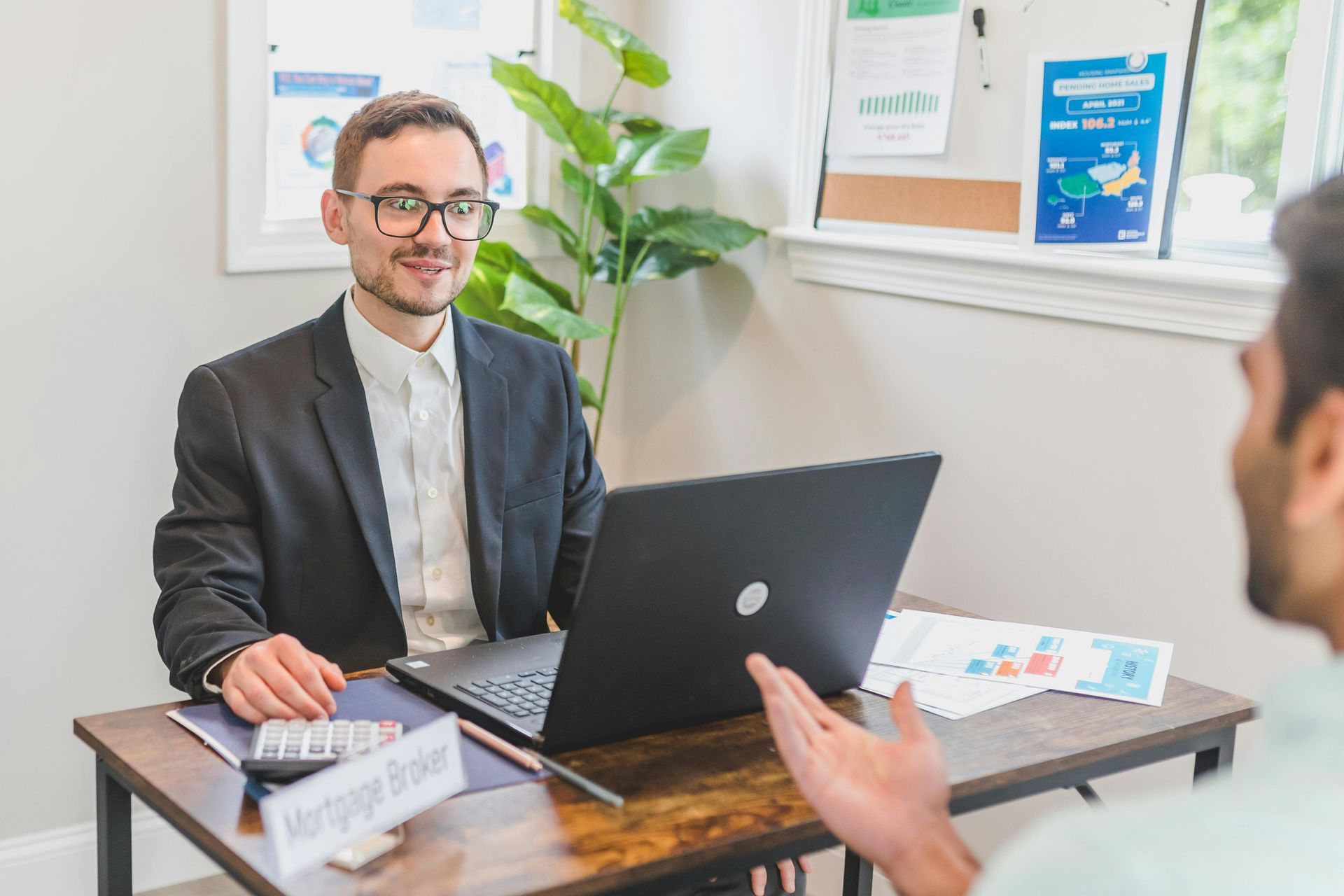 Man in suit at desk with client, discussing paperwork. Laptop open.