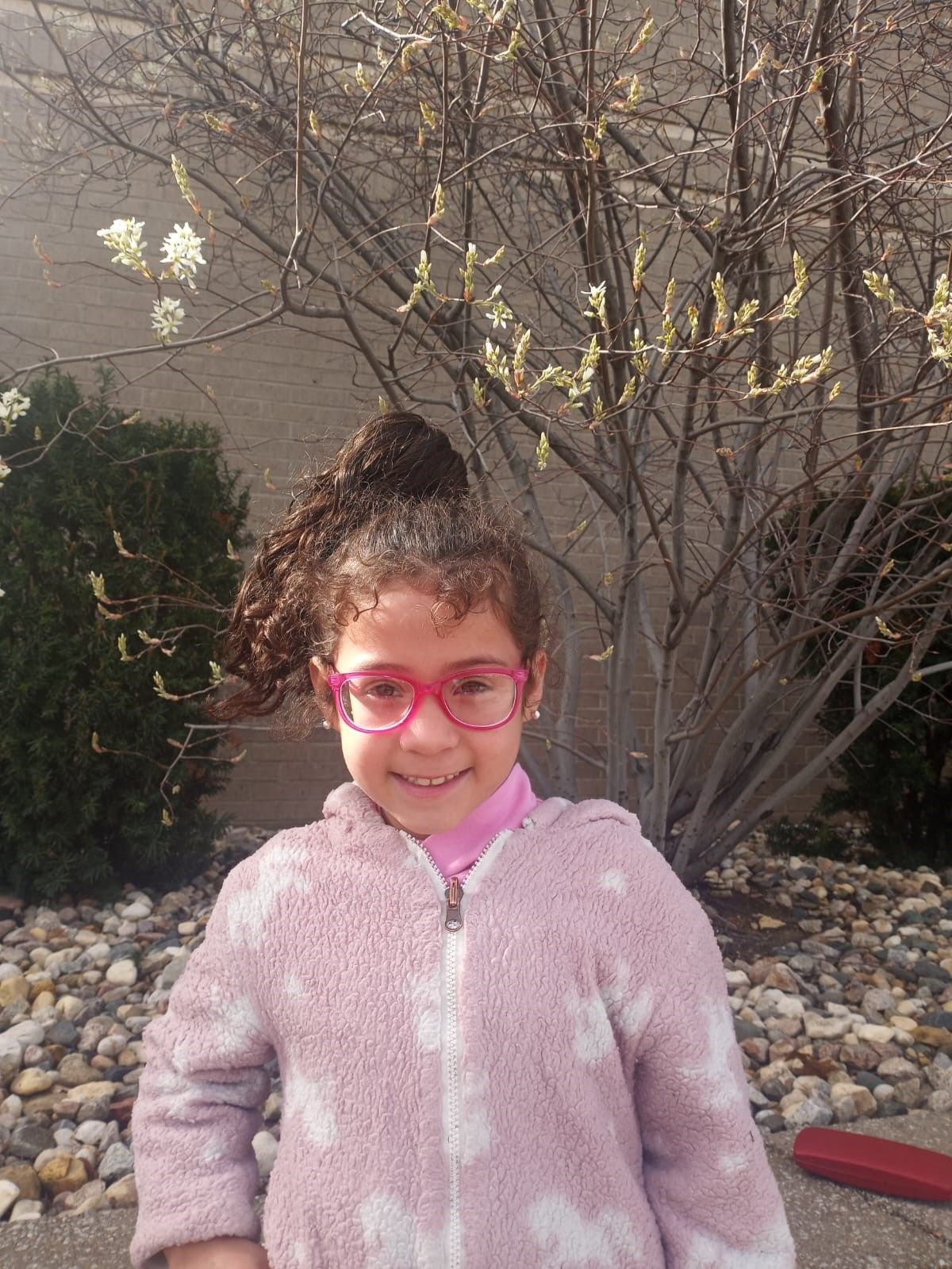A little girl wearing glasses and a pink jacket is standing in front of a tree.