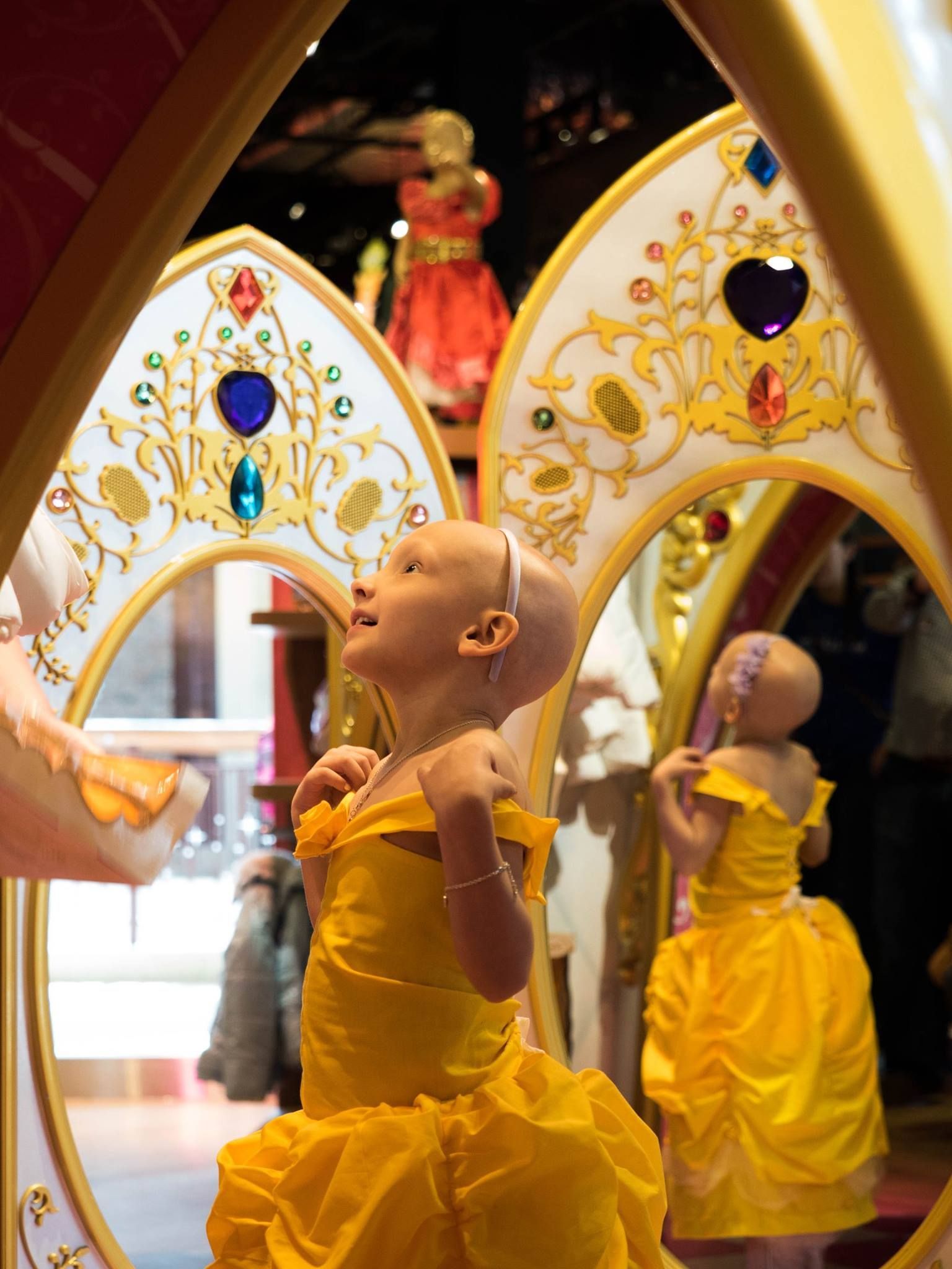 A little girl in a yellow dress is looking at herself in a mirror.