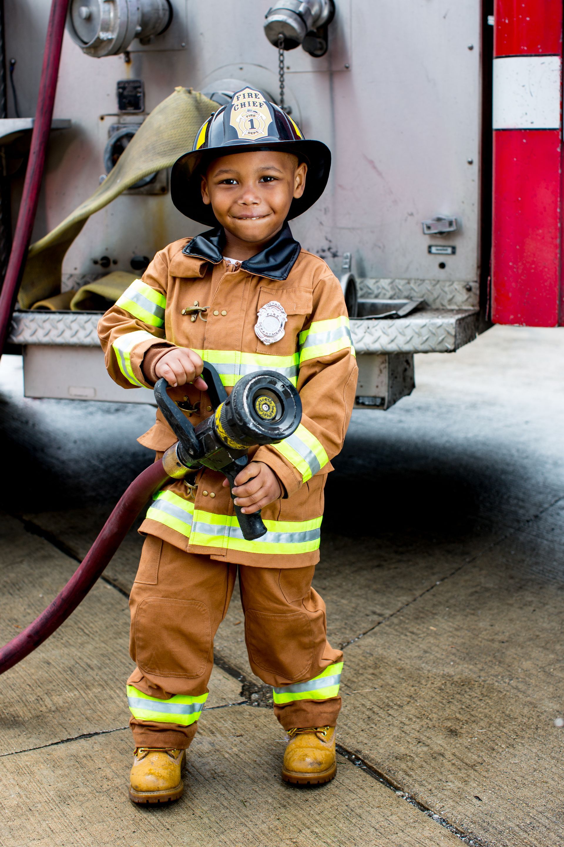 A young boy dressed as a fireman is holding a fire hose in front of a fire truck.