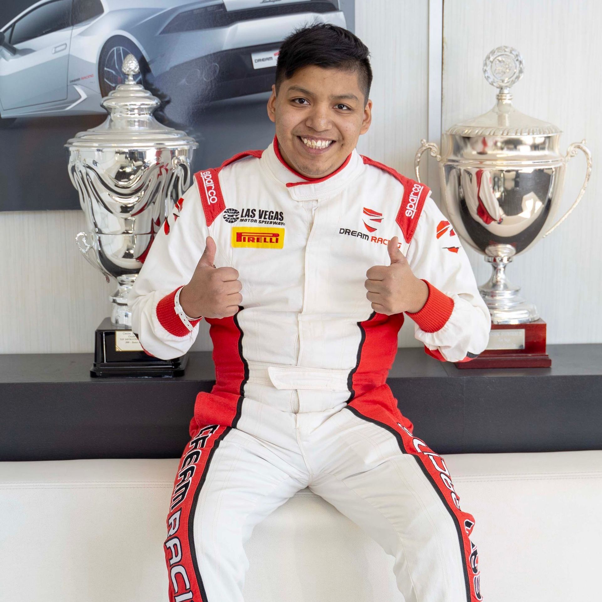 A man in a racing suit is sitting on a couch and giving a thumbs up