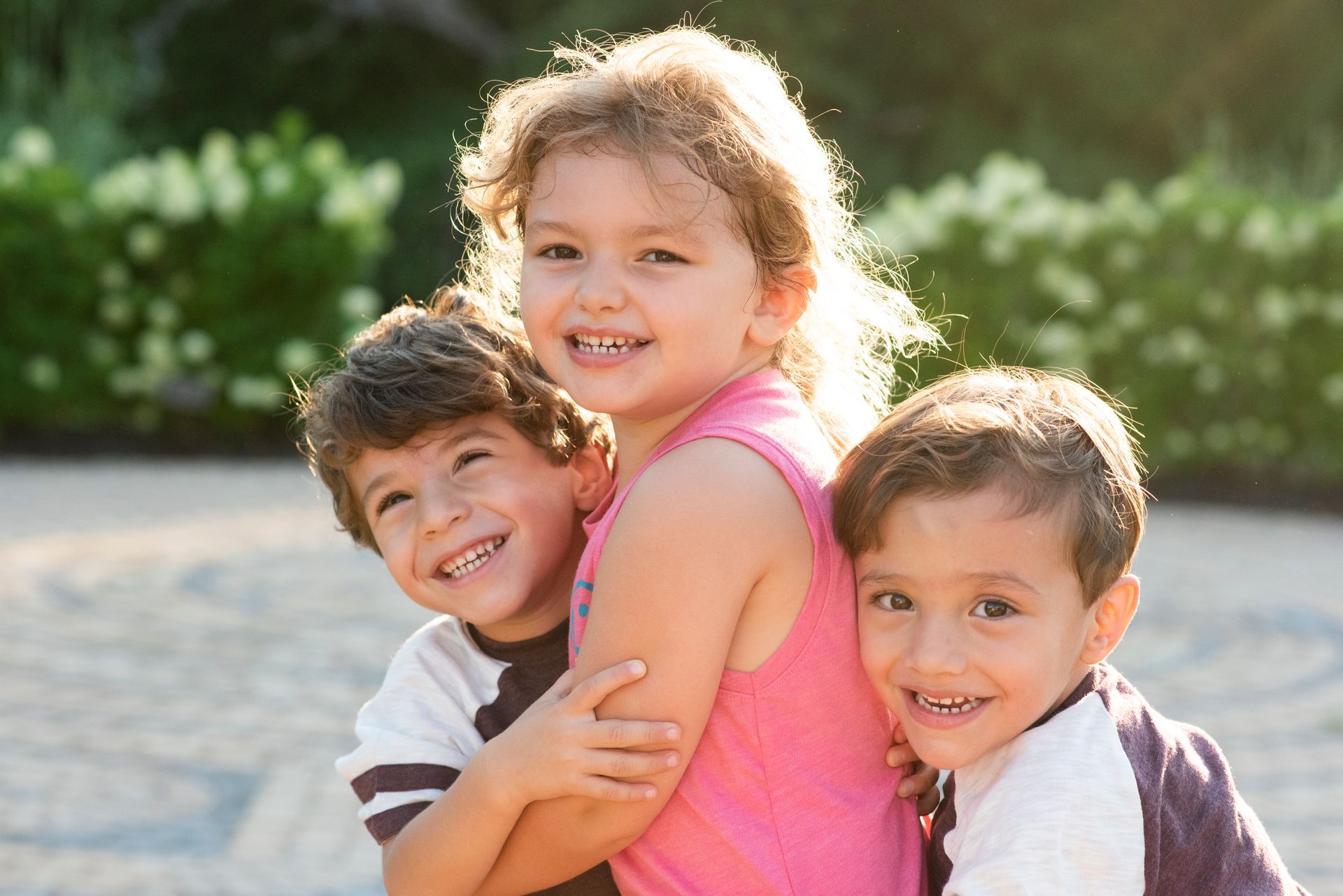 A little girl in a pink tank top is hugging two young boys