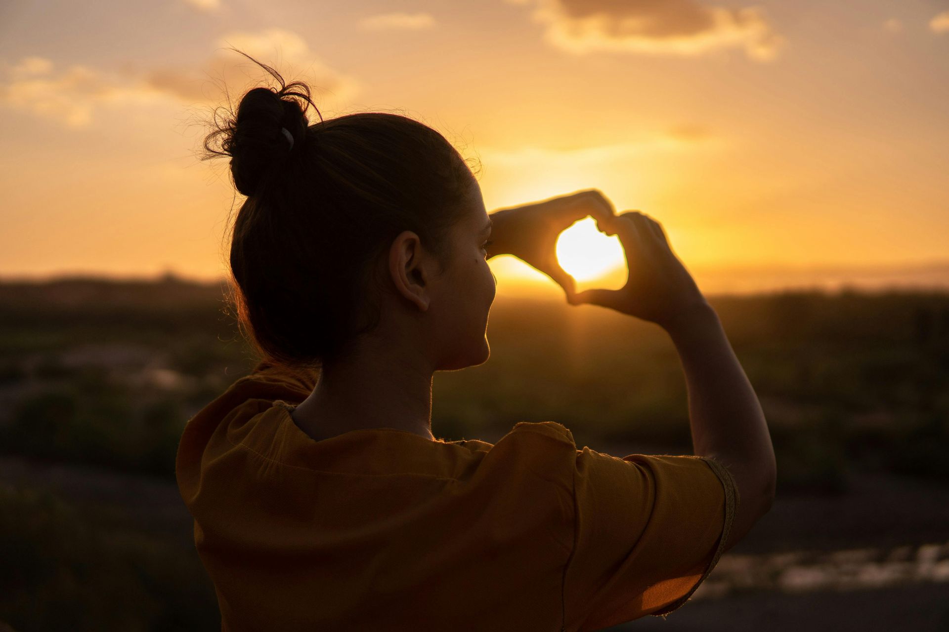 A woman is making a heart shape with her hands in front of the sun at sunset.