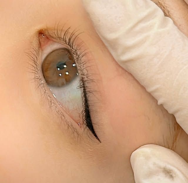 Eye with eyeliner being applied, eyelid held by gloved hand.