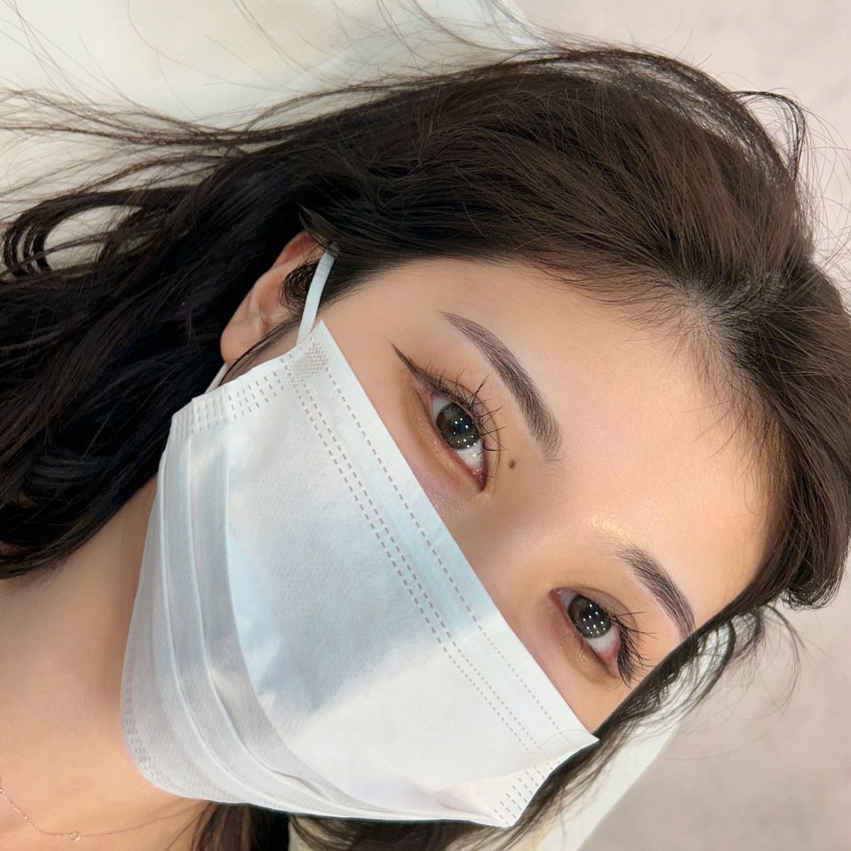 Woman wearing a white face mask, brown eyes, and dark eyebrows, lying down.