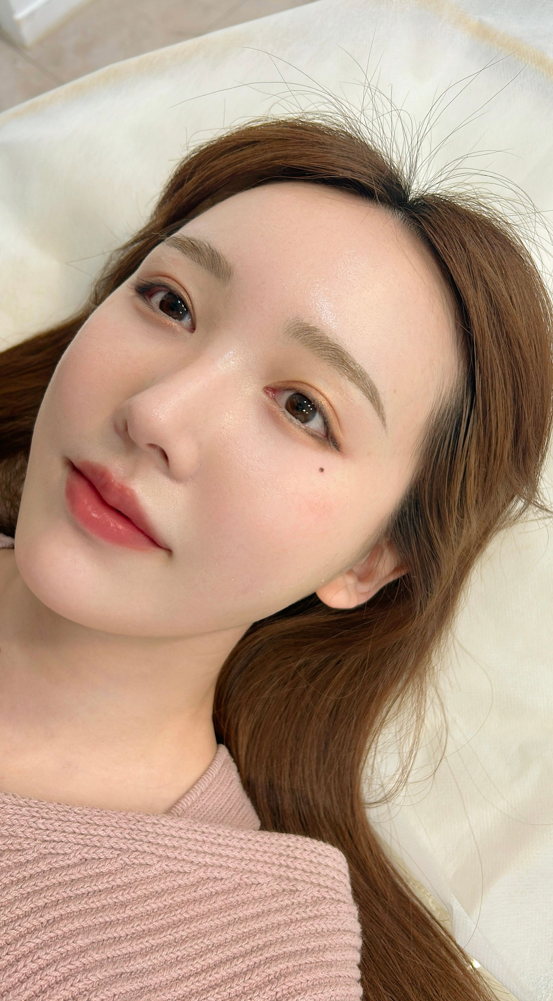 Woman with light makeup, lying down. Wearing a pink shirt. Brown hair.