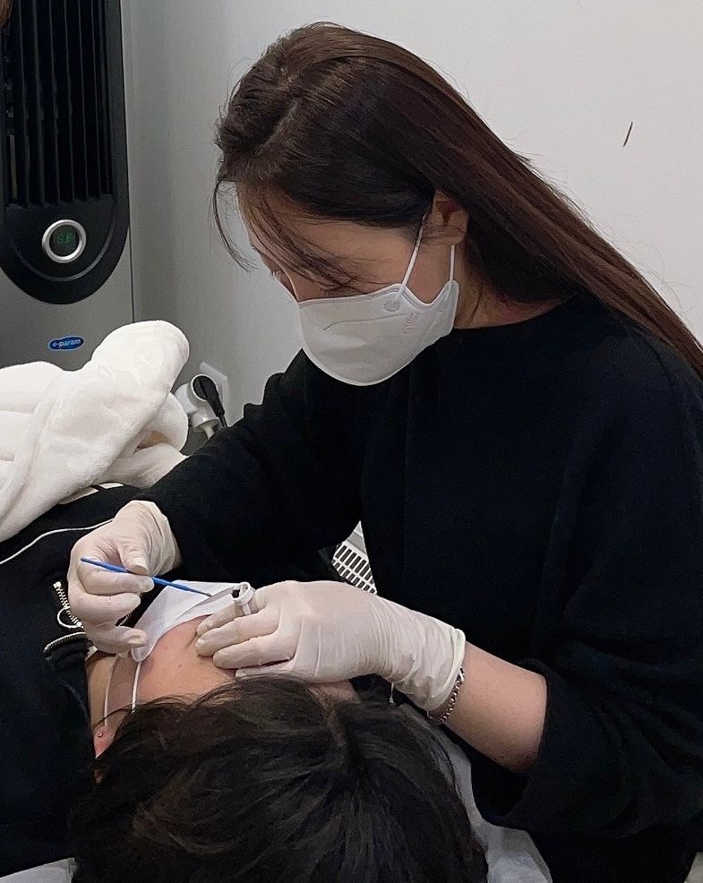 Woman in mask and gloves working on a person's eyebrow. In an office setting, using a tool.