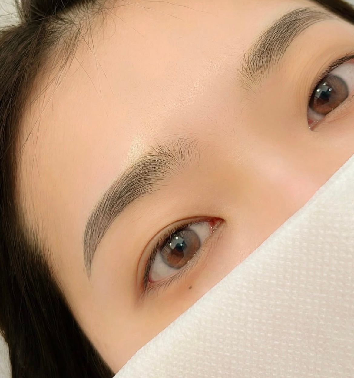 Close-up of a person's eyes and eyebrows; the person partially covered with a white cloth. Dark brown eyebrows and light brown eyes.