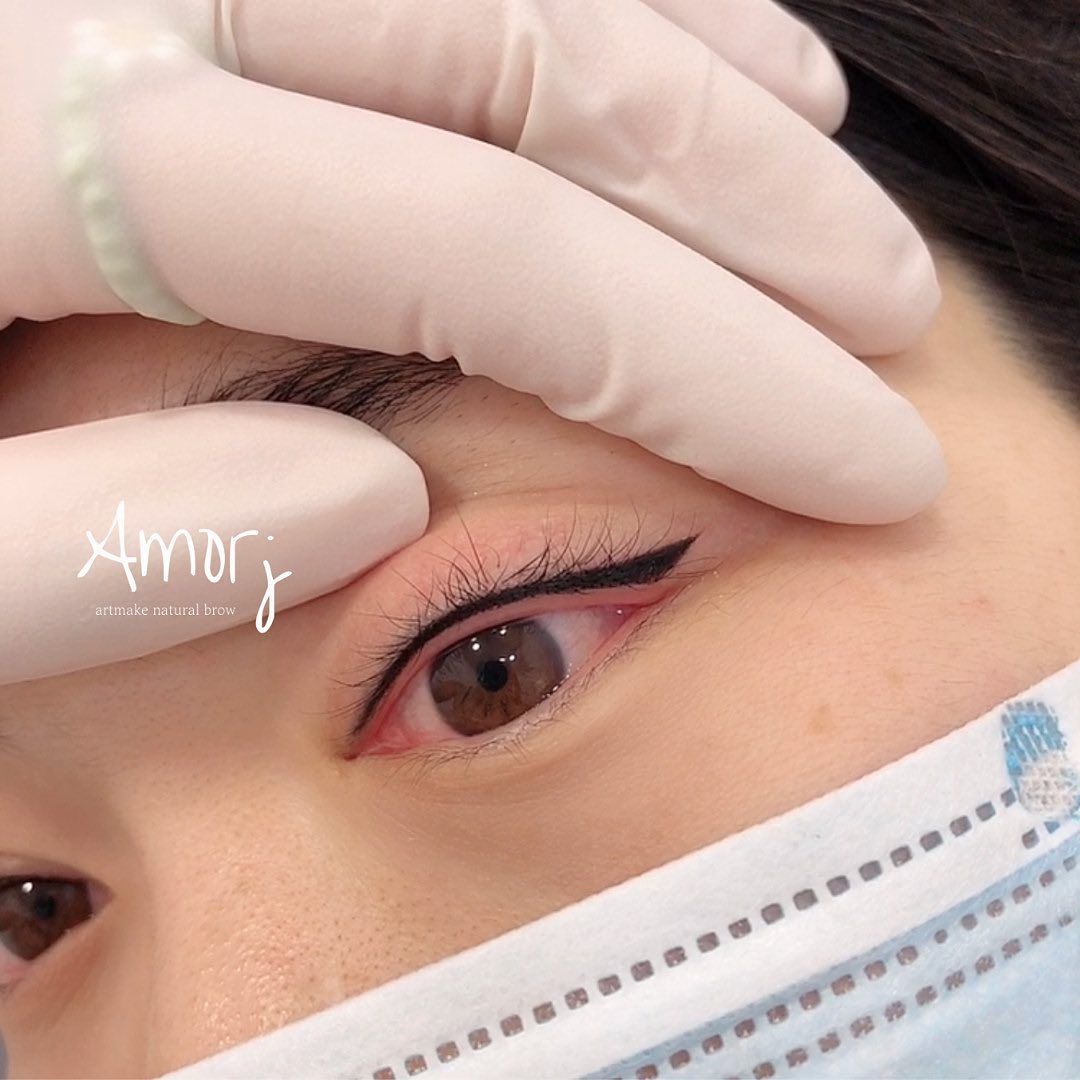 Eyeliner tattoo. Person's eye with a black winged eyeliner. Gloved hand stretches skin. Mask and glove visible.