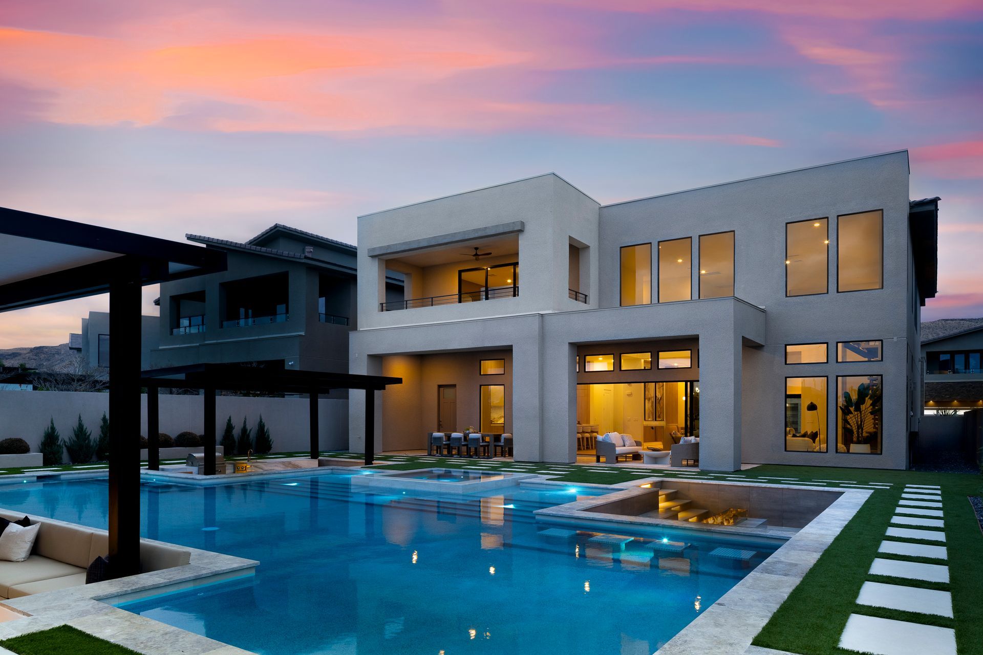 Modern home with pool at sunset. Exterior view with two-story structure, pool, spa, and pergola. Pink and blue sky.